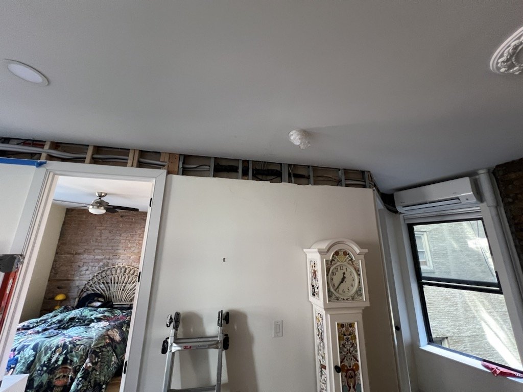 Room undergoing renovation with exposed wall studs and partially removed drywall on the ceiling, a white decorative clock, and a window with a view outside.
