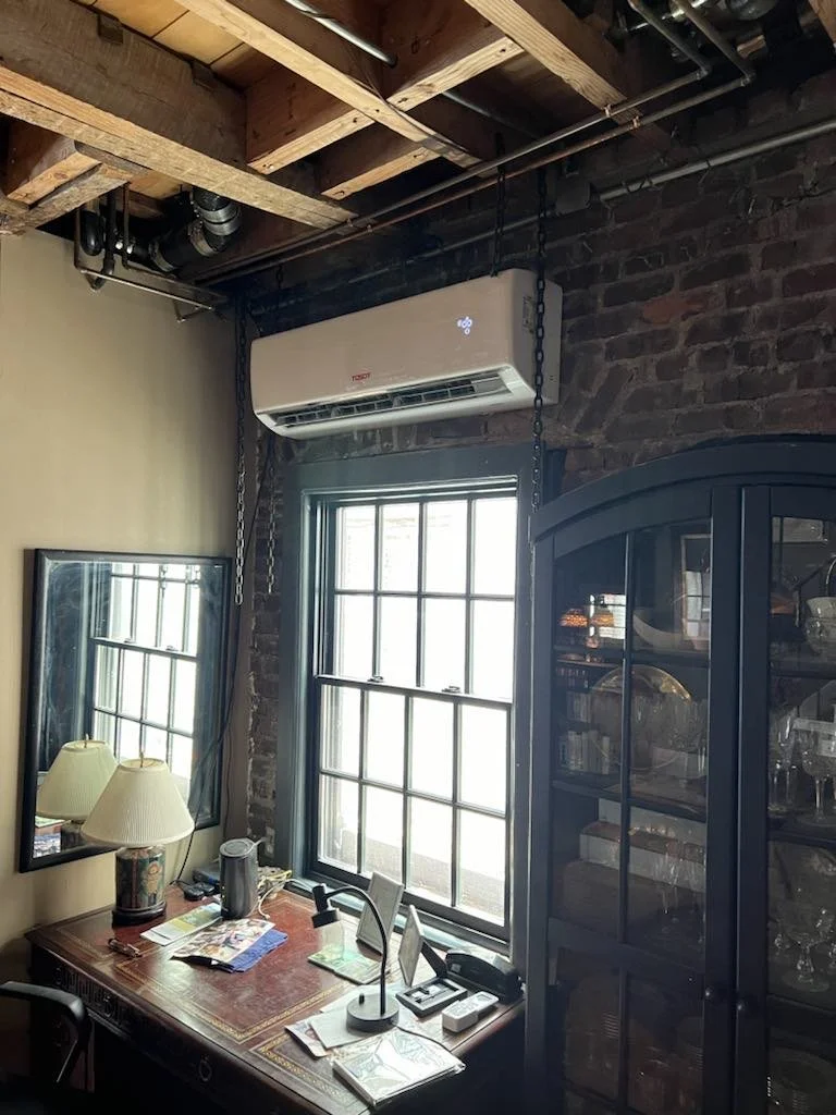 Interior view of a room with brick walls, a window, a desk with lamps and documents, a mirror, a black cabinet with glass doors, and an air conditioner mounted on the brick wall near the ceiling.
