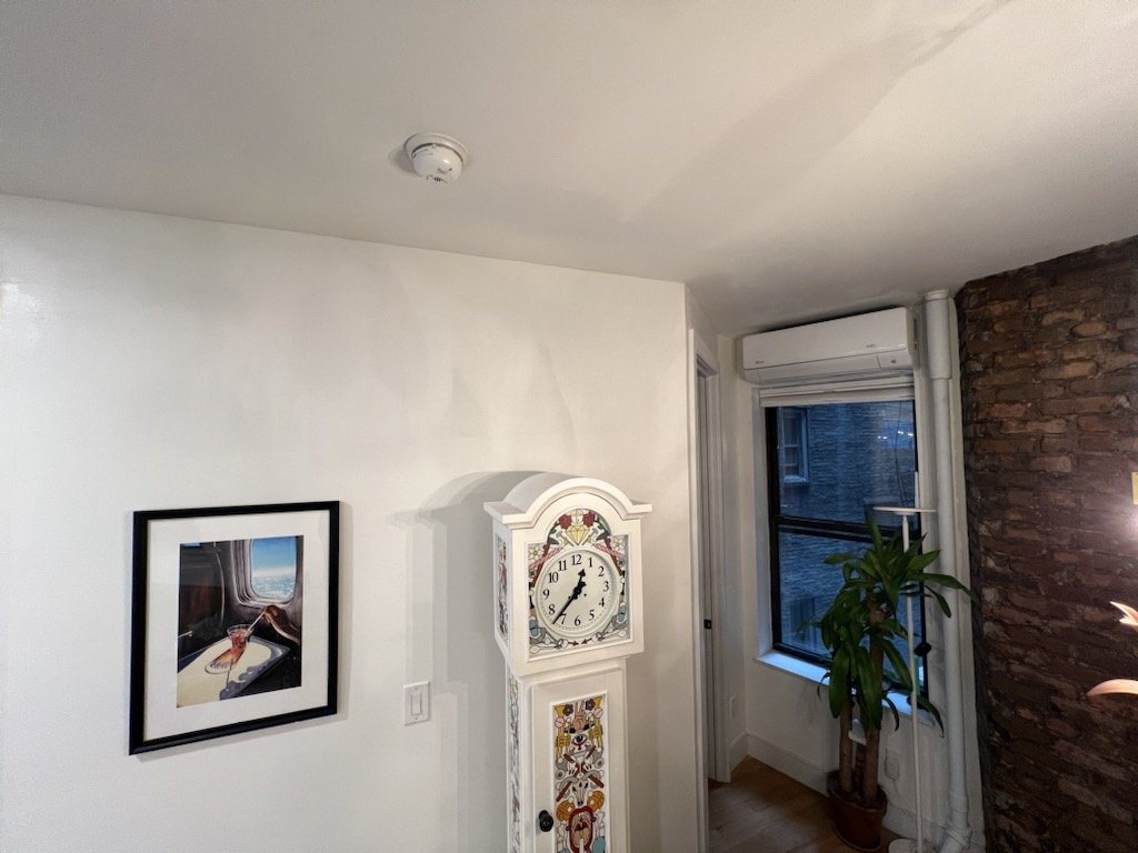 Interior of a room with a white wall, a framed picture, a white grandfather clock, a green plant, a window, and a brick accent wall.