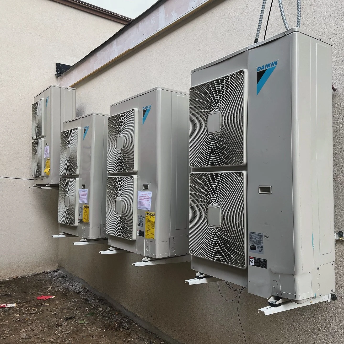 Five outdoor air conditioning units mounted on a beige wall, with a dirt ground below.
