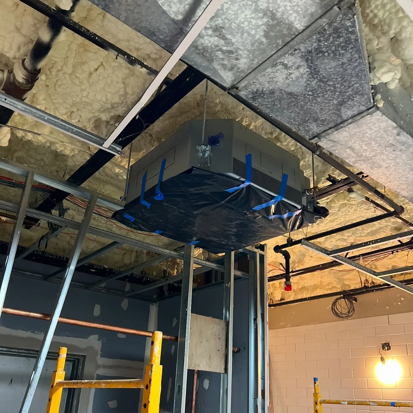 Construction site with metal framing, yellow safety railings, an HVAC unit wrapped in black plastic, and insulation foam on the ceiling.