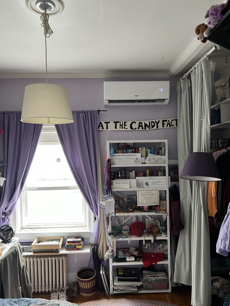 A bedroom with purple curtains, a window, and cream-colored lampshade hanging from the ceiling. A white air conditioning unit is mounted on the wall. There is a bookshelf filled with books and various items, and a sign above it that reads "AT THE CANDY FACT." Part of a closet with curtains and clothes is visible on the right.