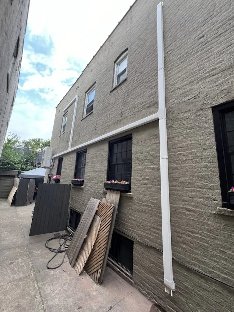 Backyard side of a brick building with four windows, three on the lower level and one on the upper level. Black window boxes with pink flowers are mounted under the lower windows. A white rain drainpipe runs down the right side of the building. Several wooden boards and garden hoses are leaning against the building's wall, and there is a gray fence and outdoor furniture in the background.