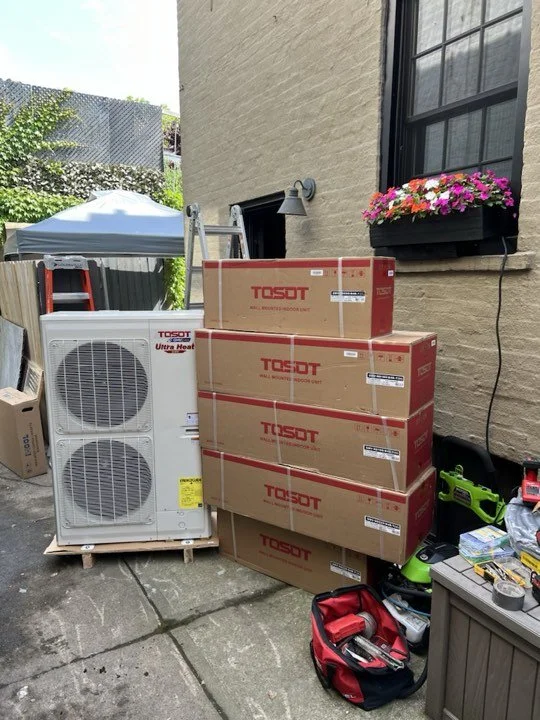 Stack of five boxed TOZOT wall-mounted roof units outside near a beige brick building with a window box of pink and purple flowers, a white air conditioning unit, a red toolbox, and various tools on the ground.