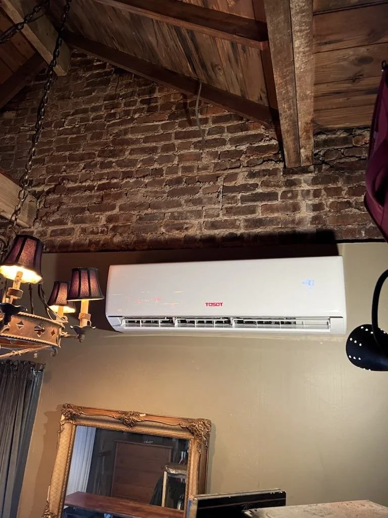 A white TOSOT air conditioning unit mounted on a beige wall inside a room with a wooden ceiling and exposed brick wall.