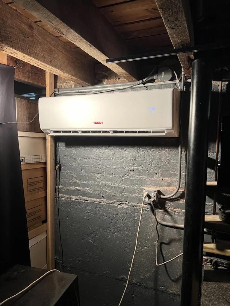 Wall-mounted air conditioner unit in a basement or utility room with exposed brick wall and shelving with storage boxes.