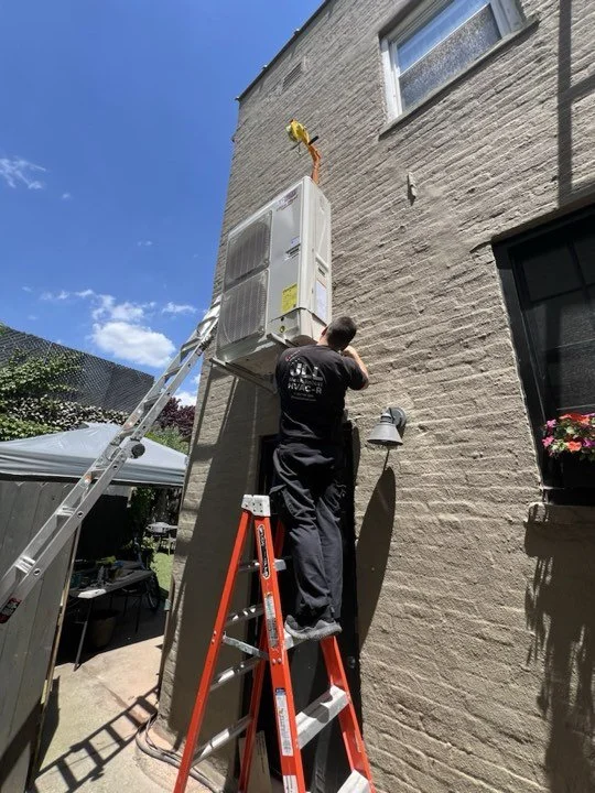 A man repairs an air conditioning unit mounted on the exterior wall of a house, standing on a red ladder on a sunny day.