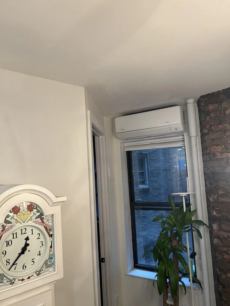 Interior corner of a room with a white clock, window with a view of gray brick building, and a tall green plant near the window. There is an air conditioning unit above the window and a brick wall on the right side.