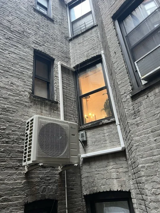 View of brick building's exterior with air conditioning unit and pipes, multiple windows reflecting interior lights, including one with a plant inside.