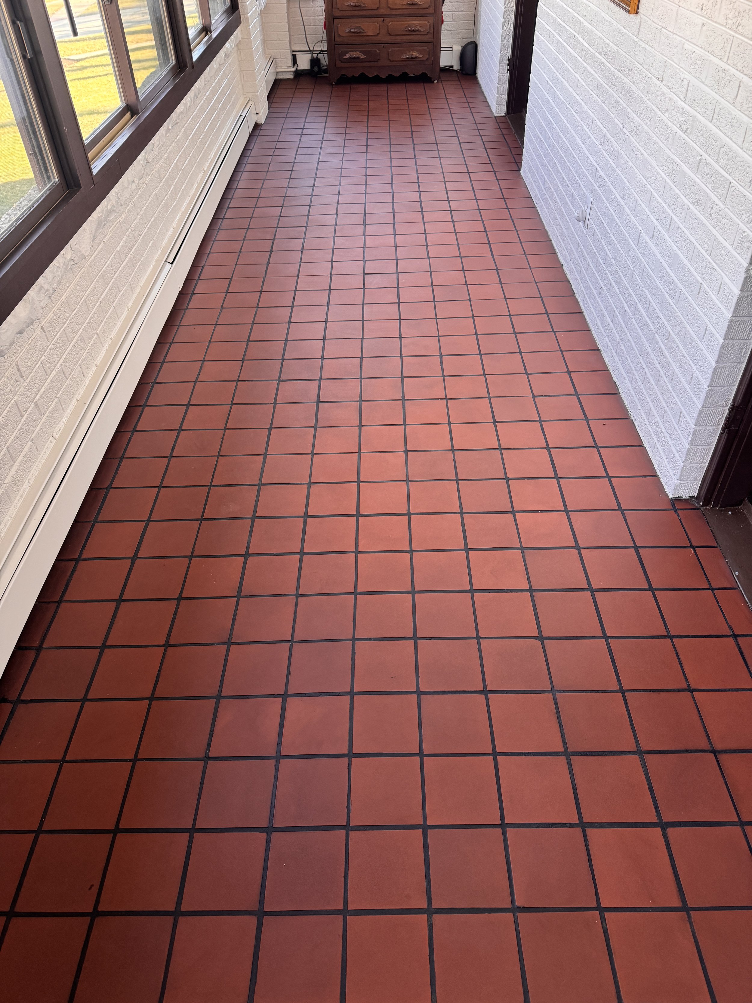 A covered porch with red square tile flooring, white brick walls, large windows, and a wooden dresser at the end.