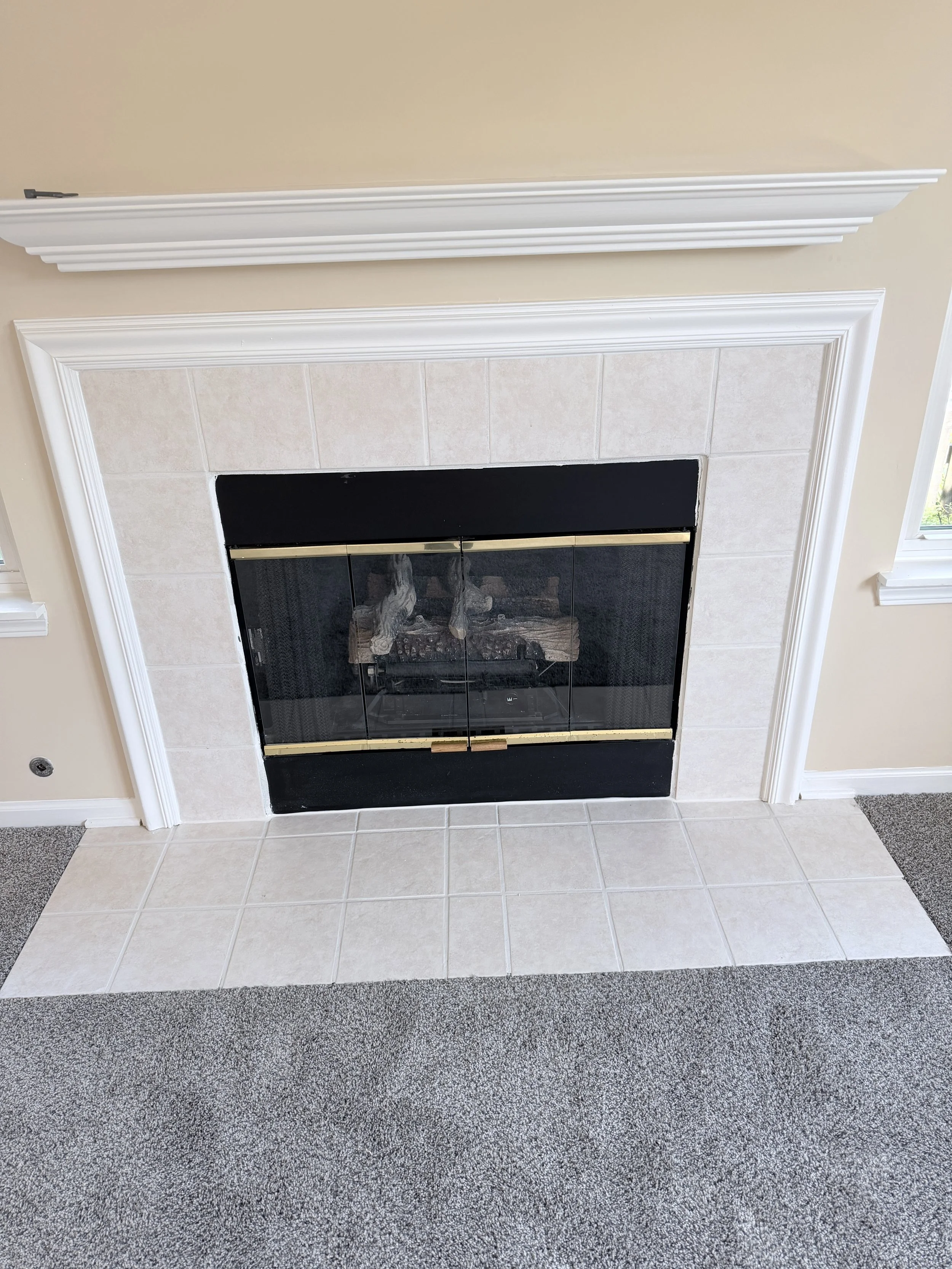 A fireplace with a white mantel, beige tile surround, and black glass doors, flanked by beige walls and windows with white trim, with a gray and beige patterned carpet in front.