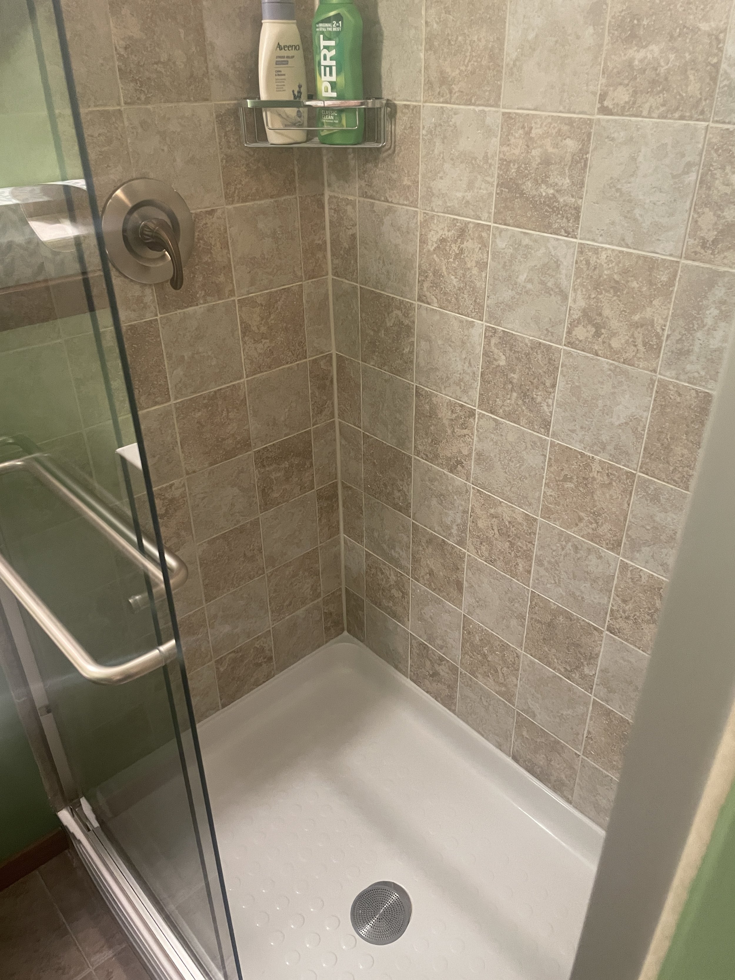 Shower with beige tiled walls, a glass door, a silver handle, a drain on the white shower floor, and a small wall-mounted metal shelf with bottles of shampoo and conditioner.
