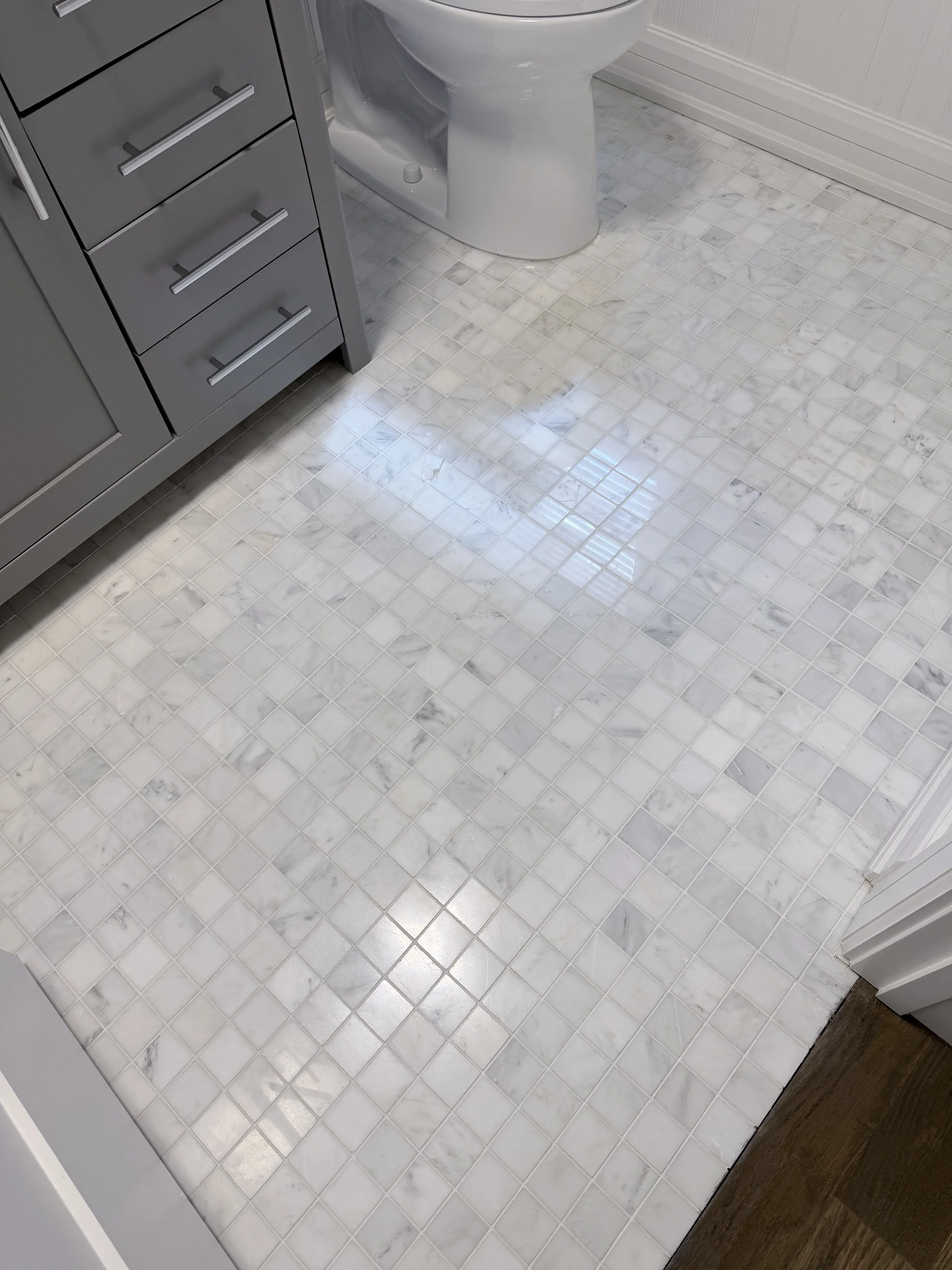 Bathroom floor with white marble tiles and a wooden floor transition at the bottom right corner. A portion of a gray cabinet with silver handles and a white toilet seat is visible.