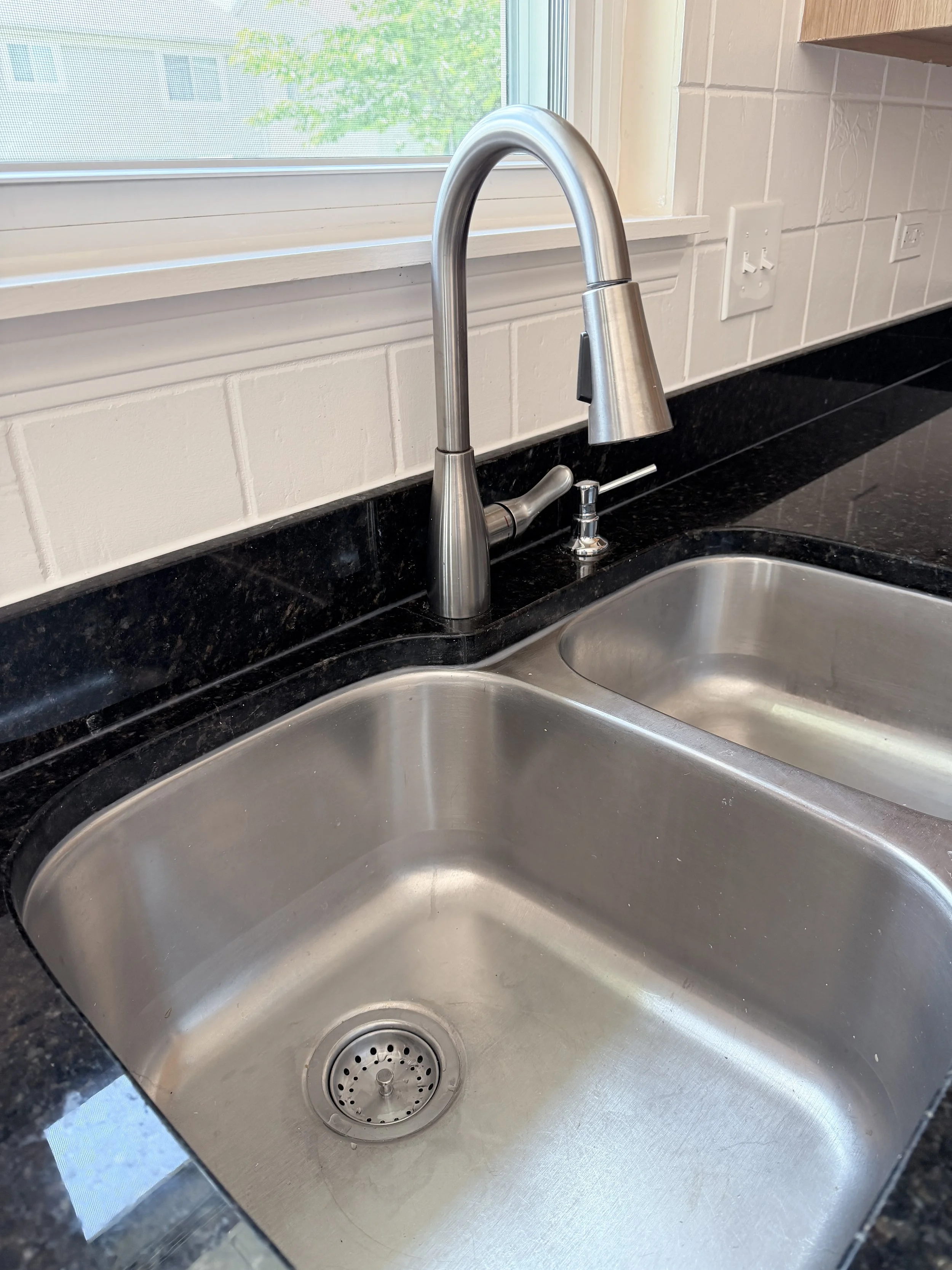 A stainless steel kitchen sink with a black granite countertop, a modern curved faucet, and a window above it showing green trees outside.