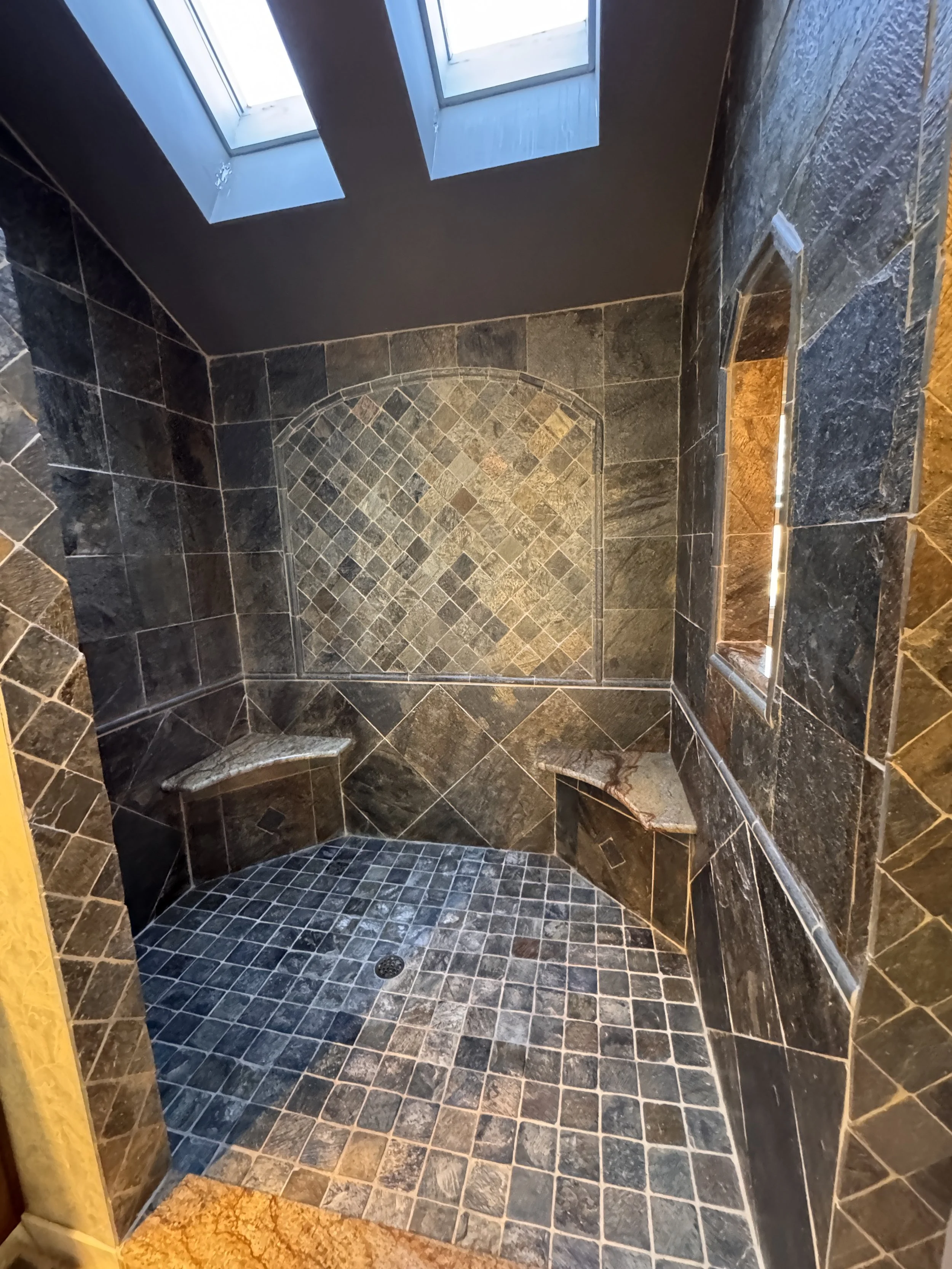 Interior view of a walk-in shower with dark slate tile walls and floor, a built-in seating bench, and two skylights providing natural lighting.