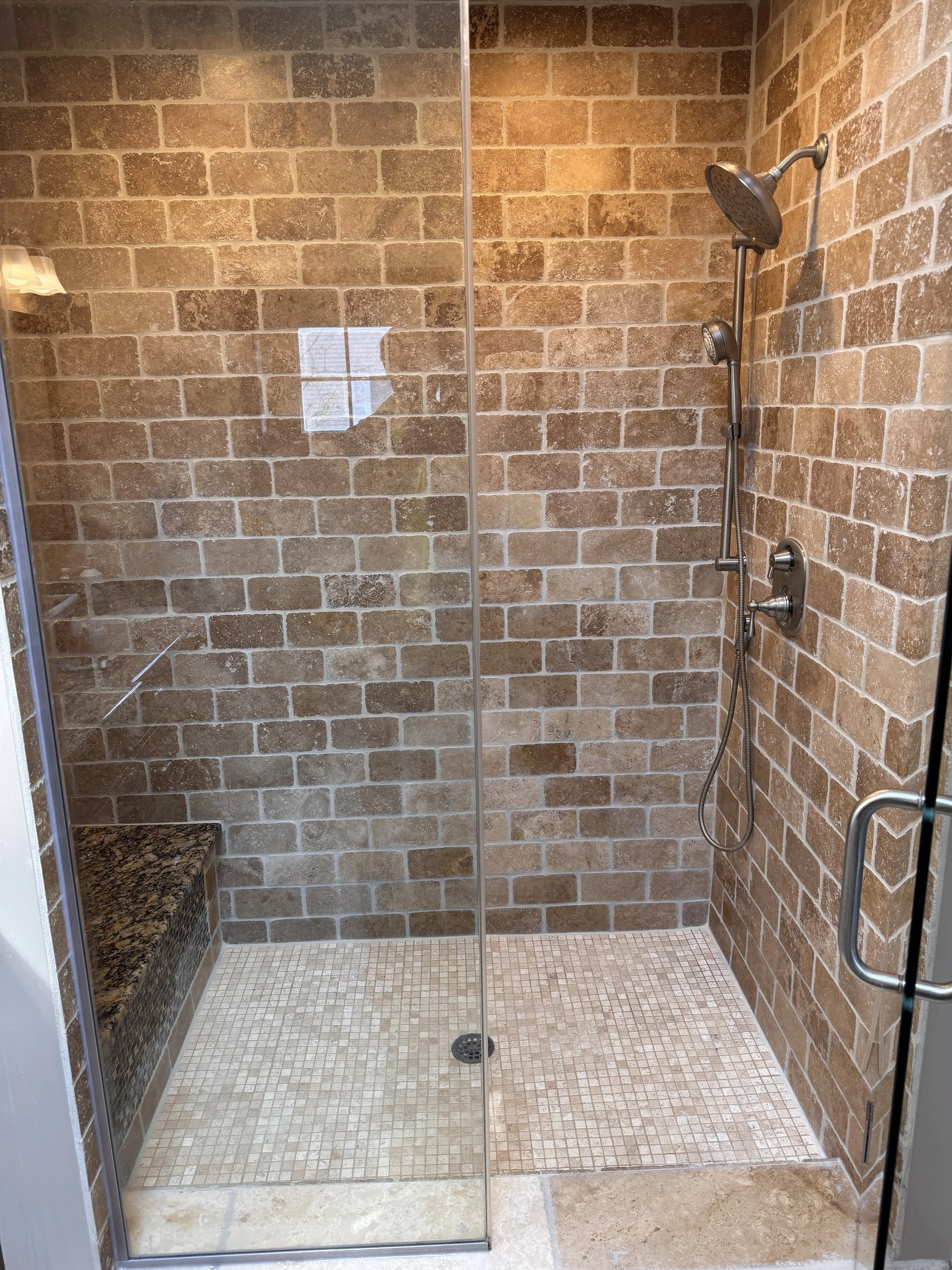 A modern walk-in shower with brick wall tiles, a granite bench on the left, and a chrome showerhead and controls on the right, with a glass door.