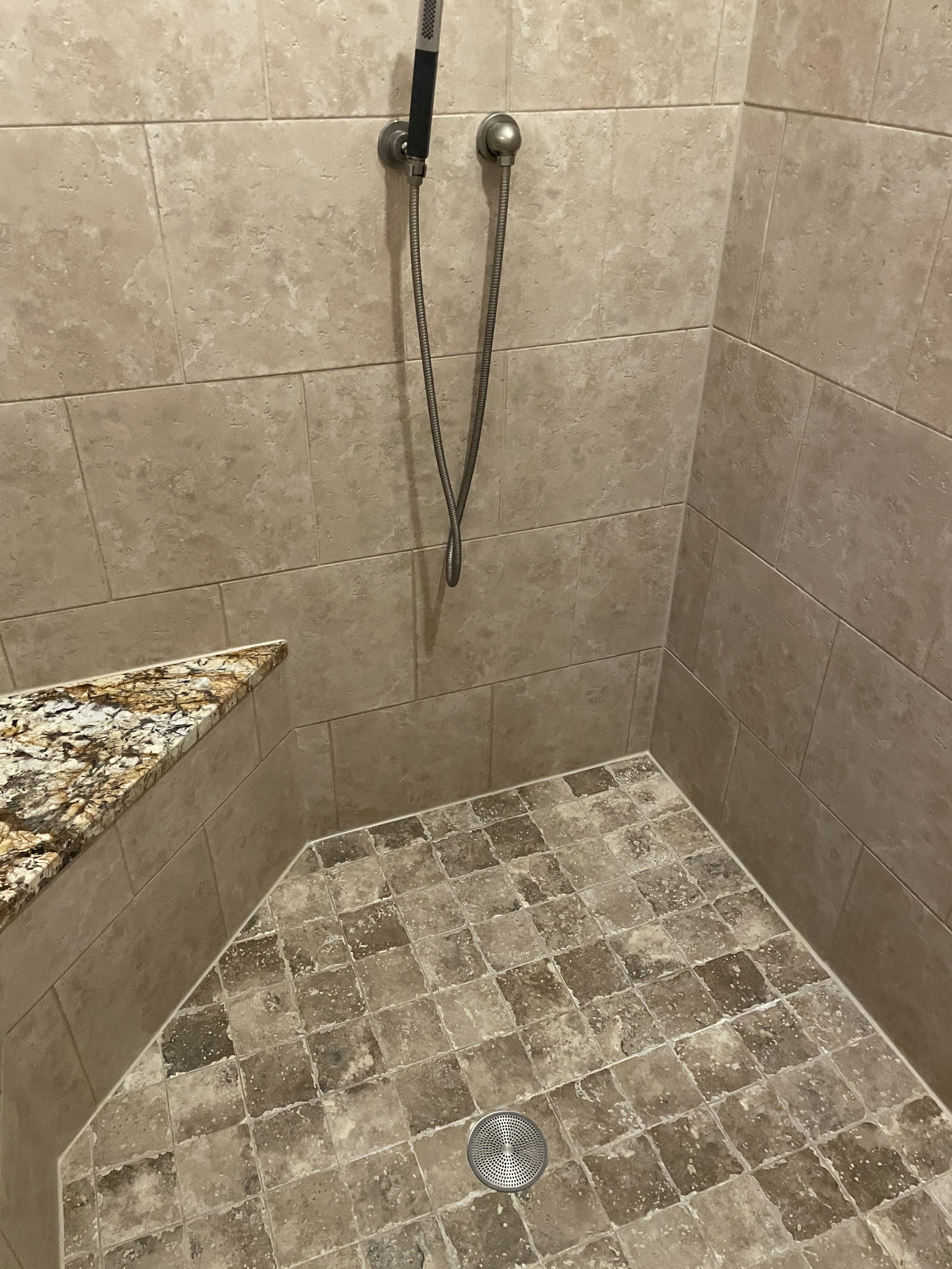 A shower stall with tiled walls and floor, a handheld showerhead mounted on the wall connected to a flexible hose, a small granite shelf, and a circular drain on the floor.