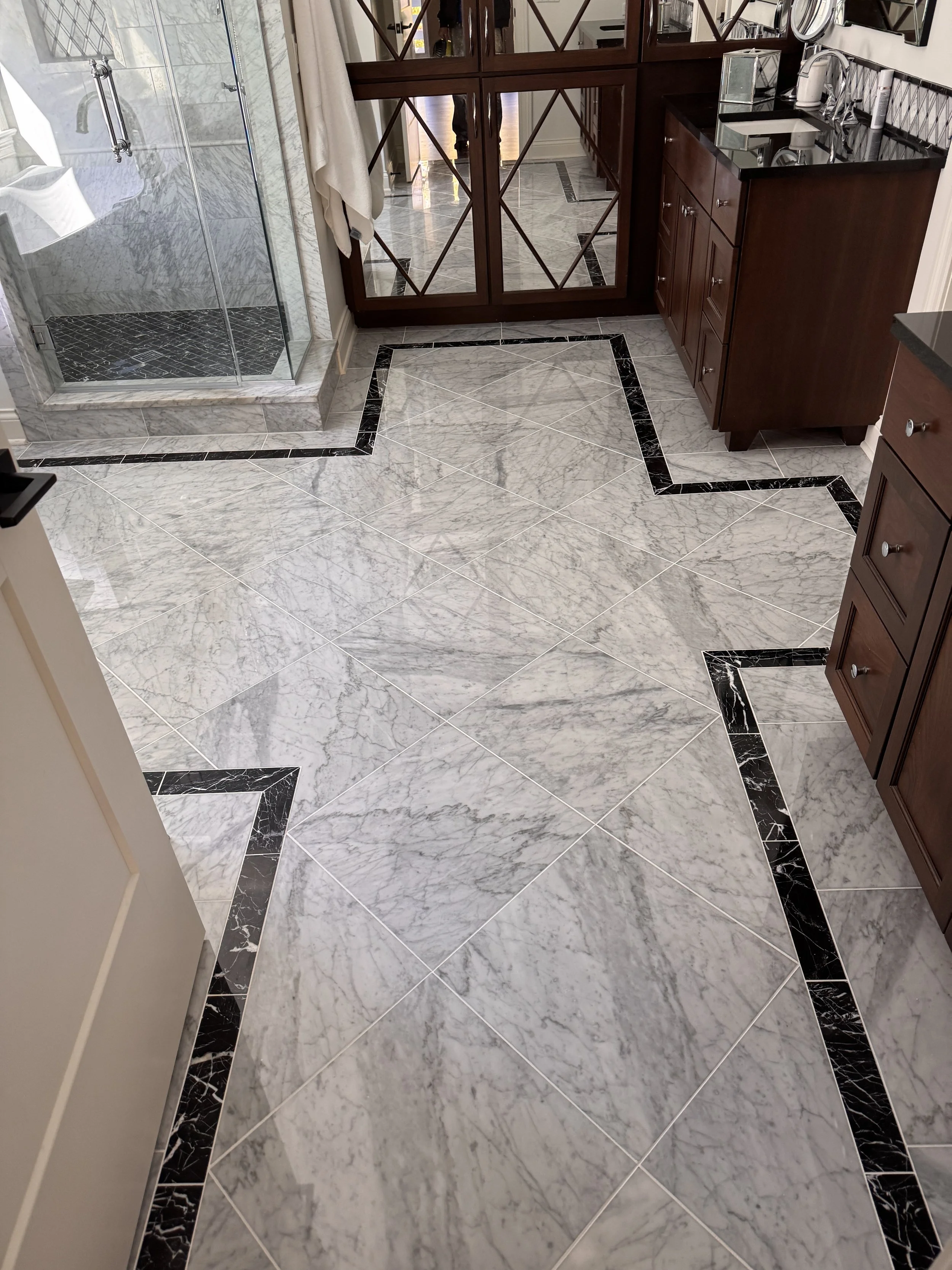 A bathroom with marble flooring and a glass shower enclosure on the left. There are wooden cabinets with black countertops and a sink on the right. A mirror and some small decorative items are visible on the wall above the cabinets. There is a person
