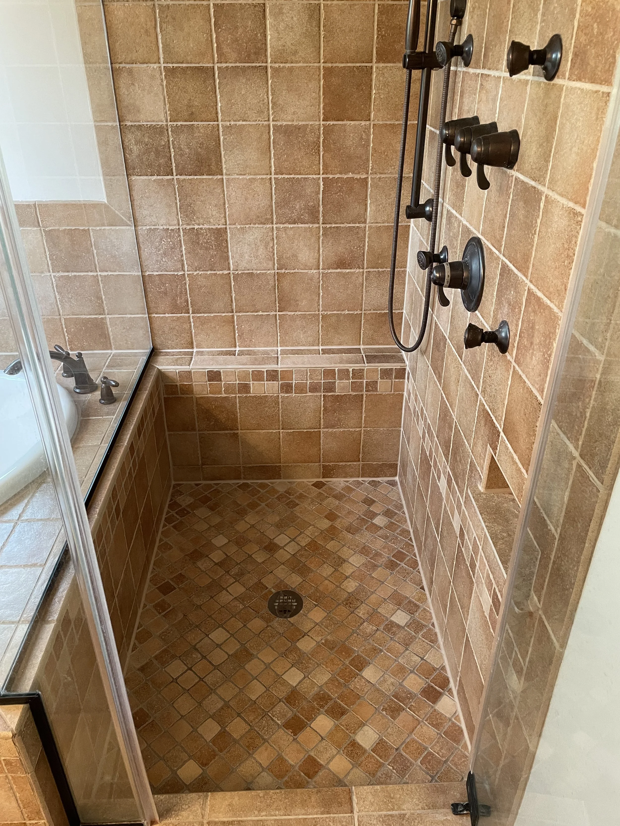 A walk-in shower with brown tiled walls and small square tile floor. It features a round floor drain, a mounted handheld showerhead, and multiple black knobs for water control.