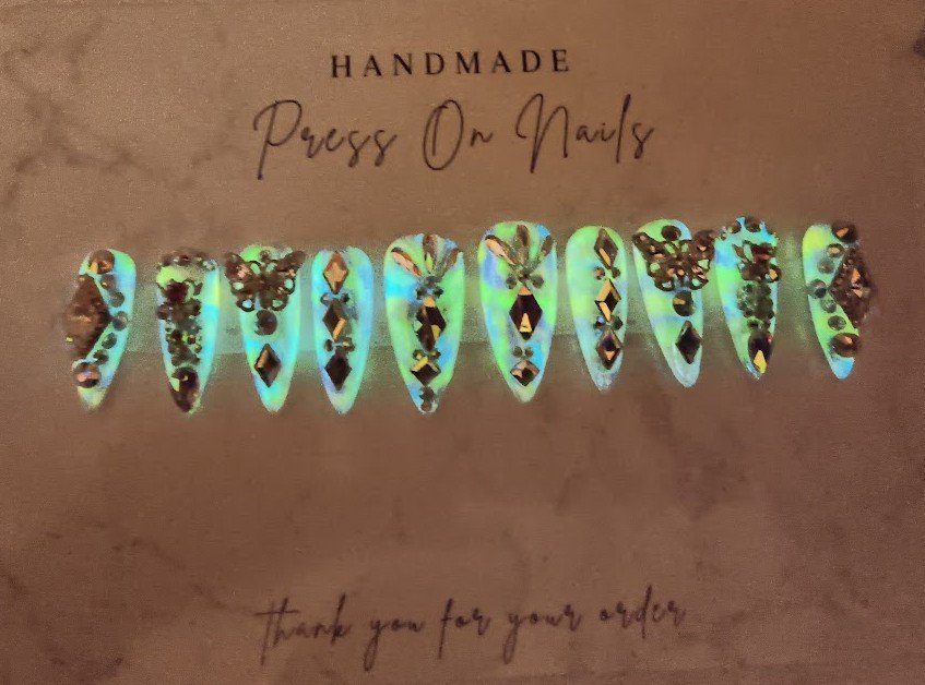 A set of iridescent, decorated press-on nails with black embellishments arranged in a row on a brown card. The card reads "Handmade Press On Nails" at the top and "thank you for your order" at the bottom.