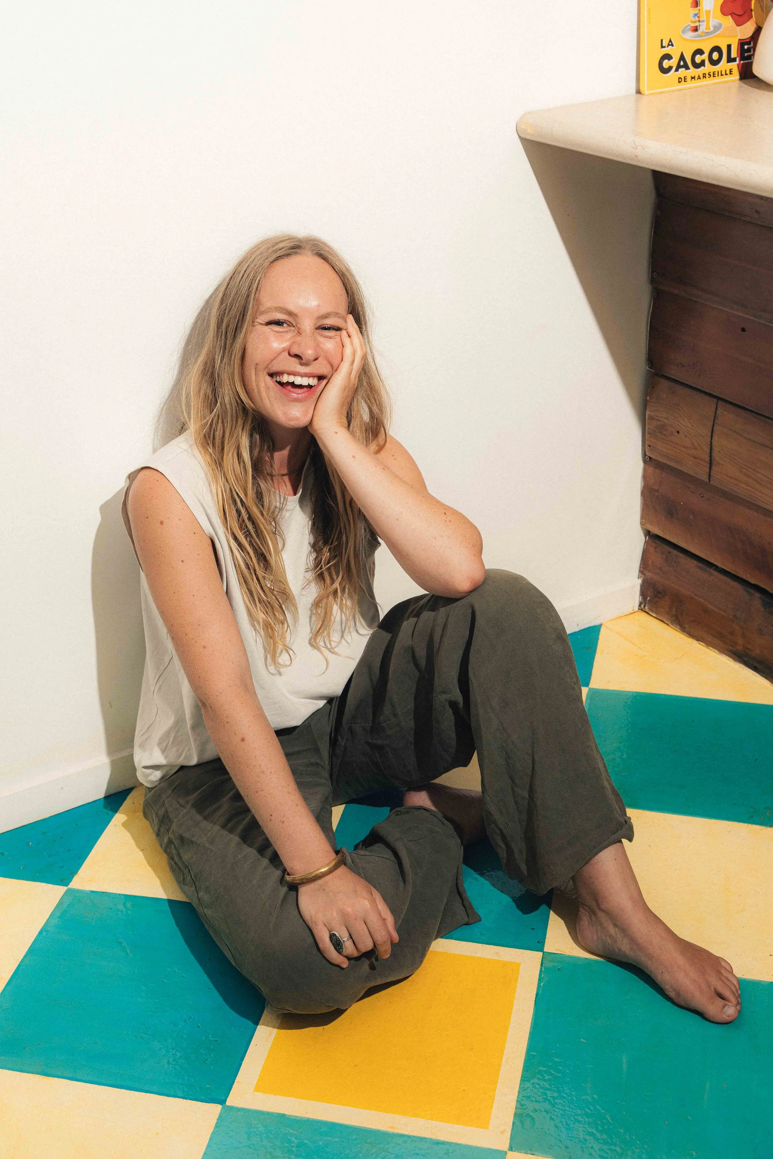 A woman with long blonde hair smiling and sitting on colorful tiled floor. She is wearing a sleeveless top and dark pants, with her hand resting on her cheek.