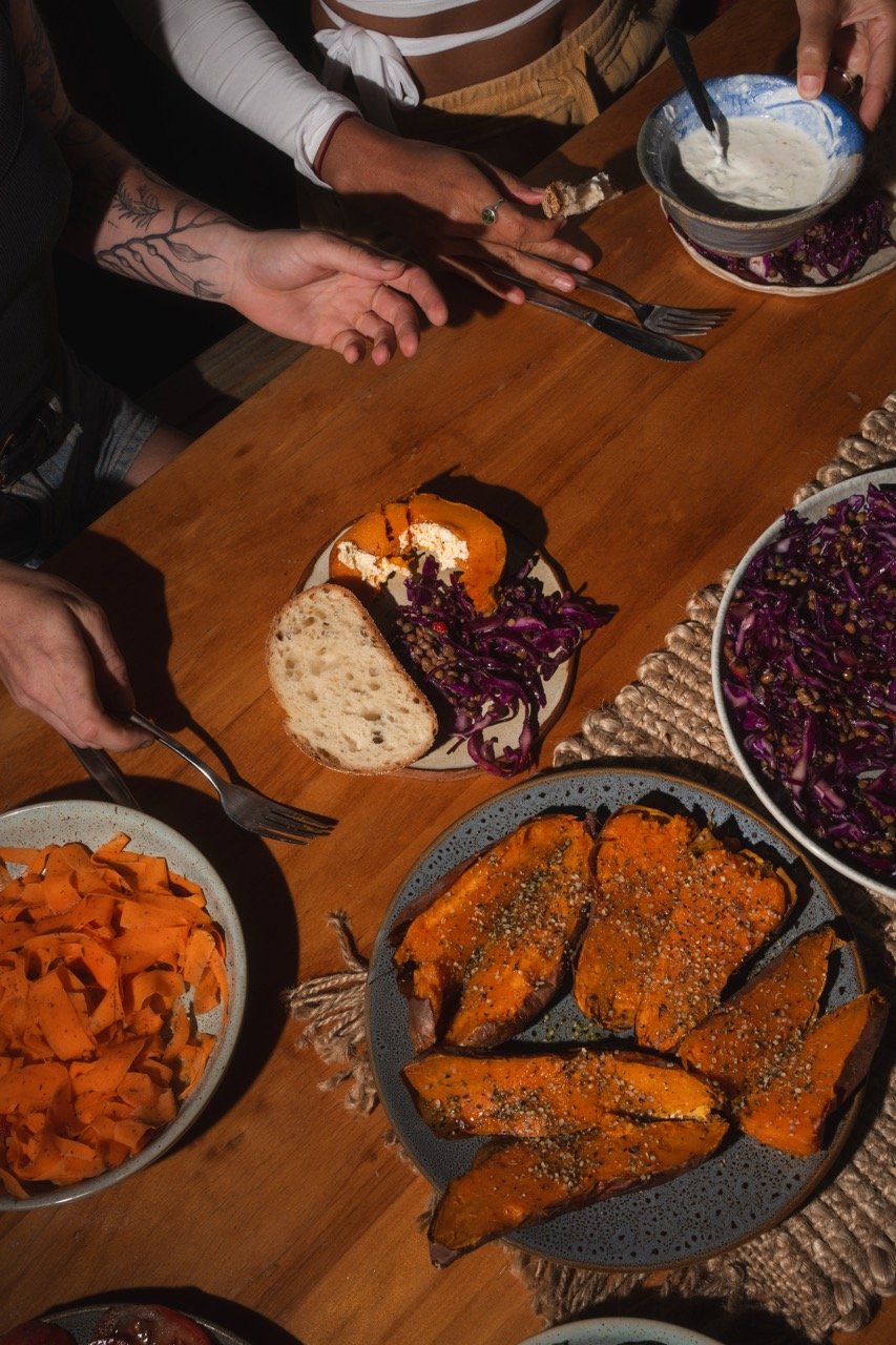 Table with various dishes including roasted sweet potatoes, a bowl of mashed potatoes, a bowl of purple cabbage slaw, and a slice of bread with purple coleslaw, with people reaching for food.