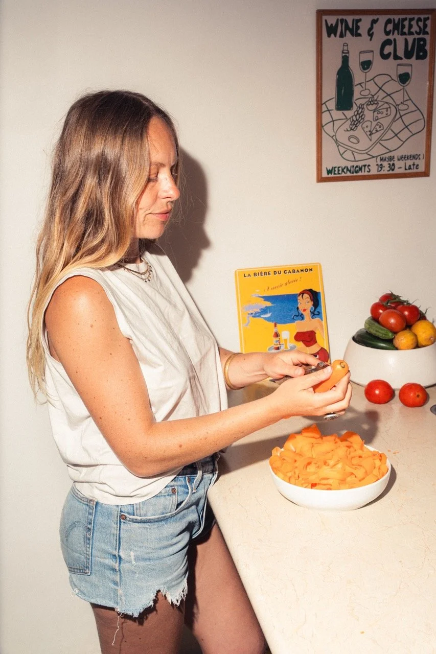 A woman with long, wavy blonde hair wearing a white sleeveless top and denim shorts peeling a carrot in a kitchen. There is a bowl of sliced carrots on the counter, a bowl with tomatoes and cucumbers, and a framed sign on the wall that says 'Wine & Cheese Club.', and a bowl of fresh tomatoes and cucumbers is on the counter.