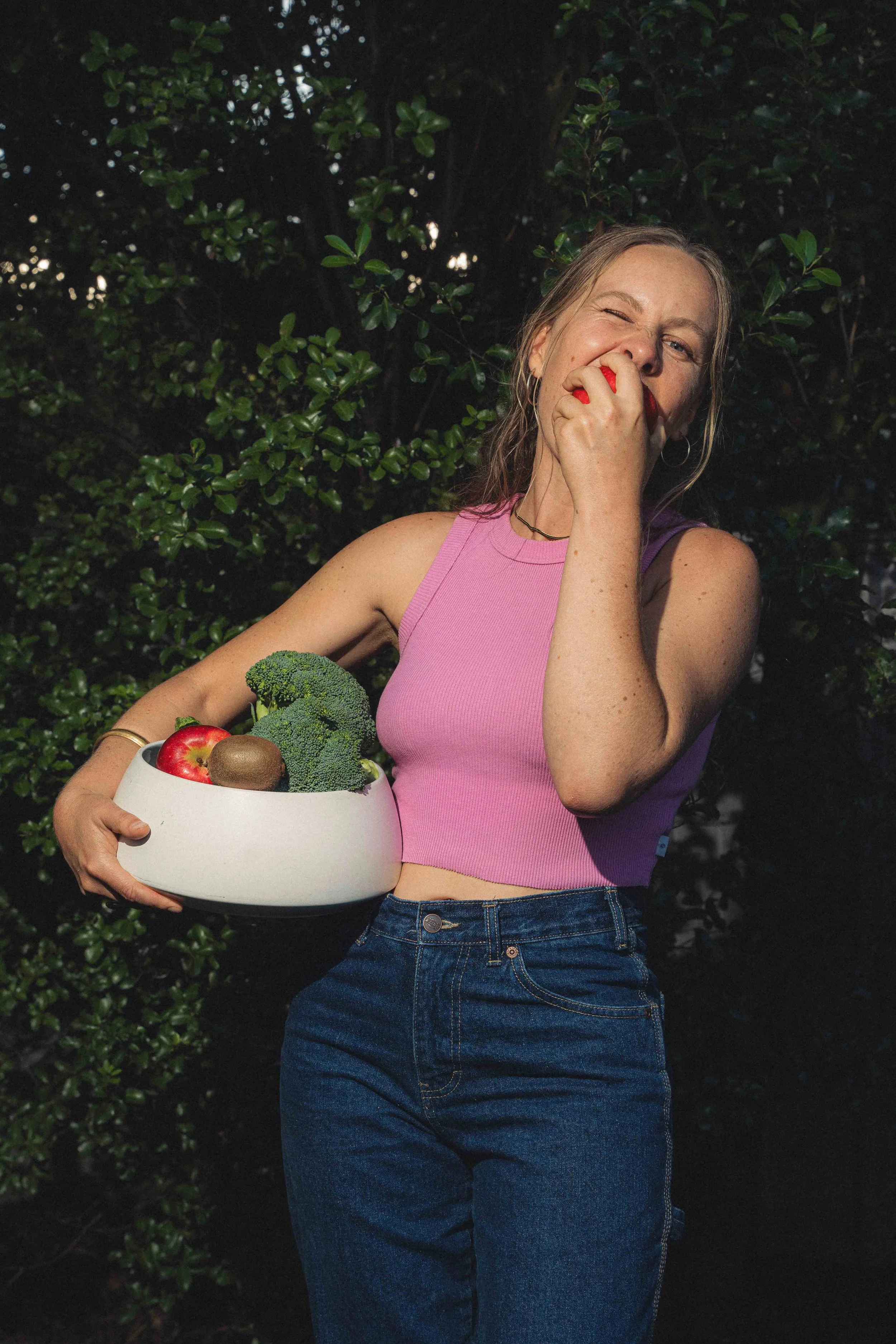 A woman dressed in a pink sleeveless top and blue jeans is standing outdoors, holding a white bowl filled with a broccoli floret, an apple, and a kiwi. She is smiling and bitting into an apple, with a background of dark green foliage.