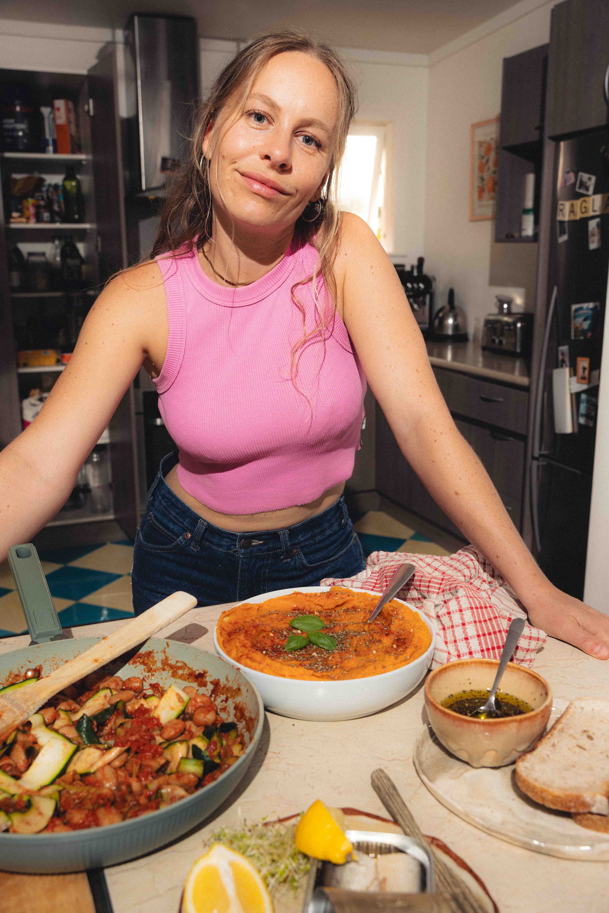 A woman in a pink sleeveless top stands in front of a table with various dishes, including a bowl of mashed carrot, vegetables, and sauces, in a kitchen setting with dark cabinets and a refrigerator.
