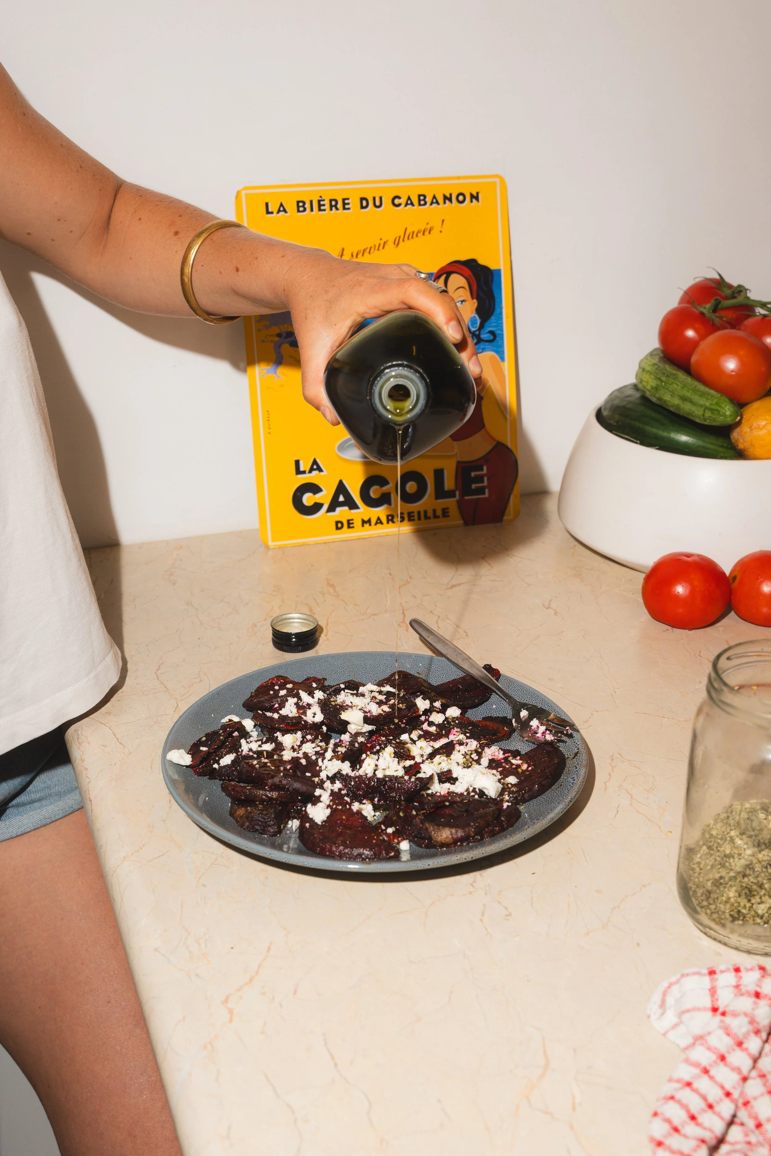 A hand pouring extra virgin olive oil onto a plate of cooked beetroot slices topped with feta, with a yellow vintage advertisement poster and a bowl of tomatoes and cucumbers on the countertop.