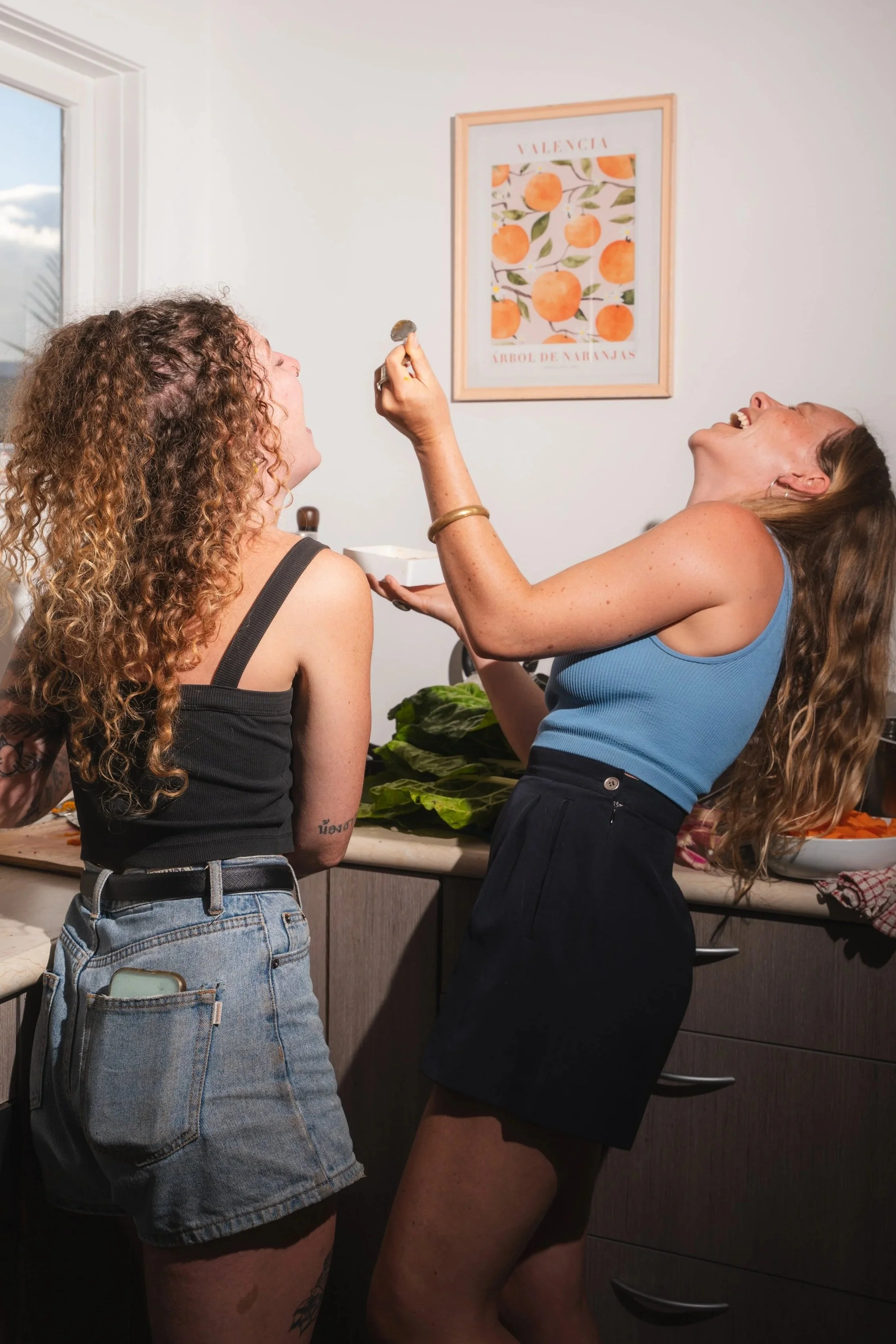 Two women laughing, one with curly hair and tattoo, wearing a black tank top and denim shorts, and the other with long straight hair, wearing a blue tank top and black shorts, standing in a kitchen near a counter with green vegetables, with a window and a framed picture of oranges on the wall in the background.