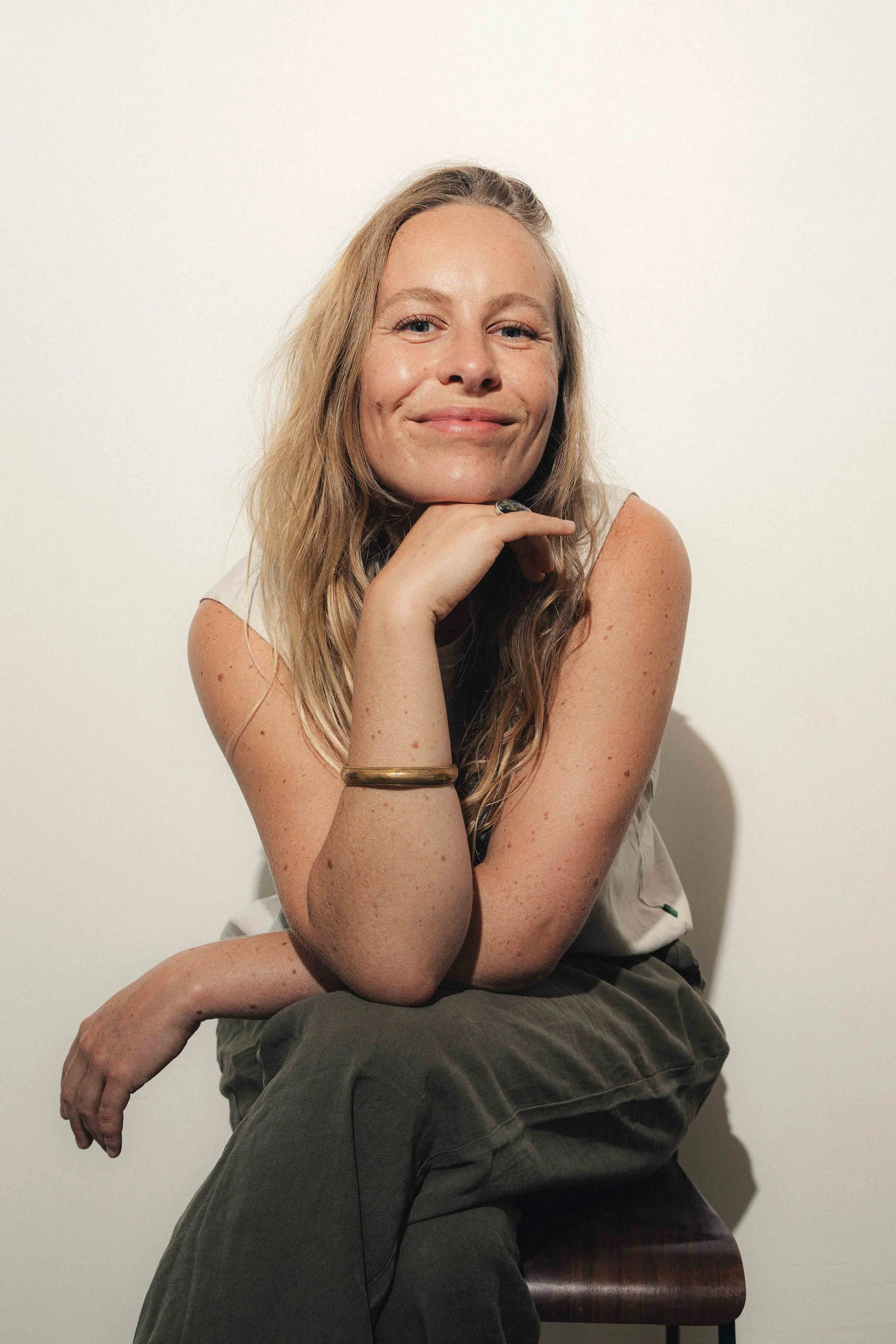 A woman with long blond hair, light skin, and freckles, smiling with her eyes slightly squinted and resting her chin on her hand. She wears a sleeveless top, a bracelet, and a ring, and is seated on a chair with a white background.