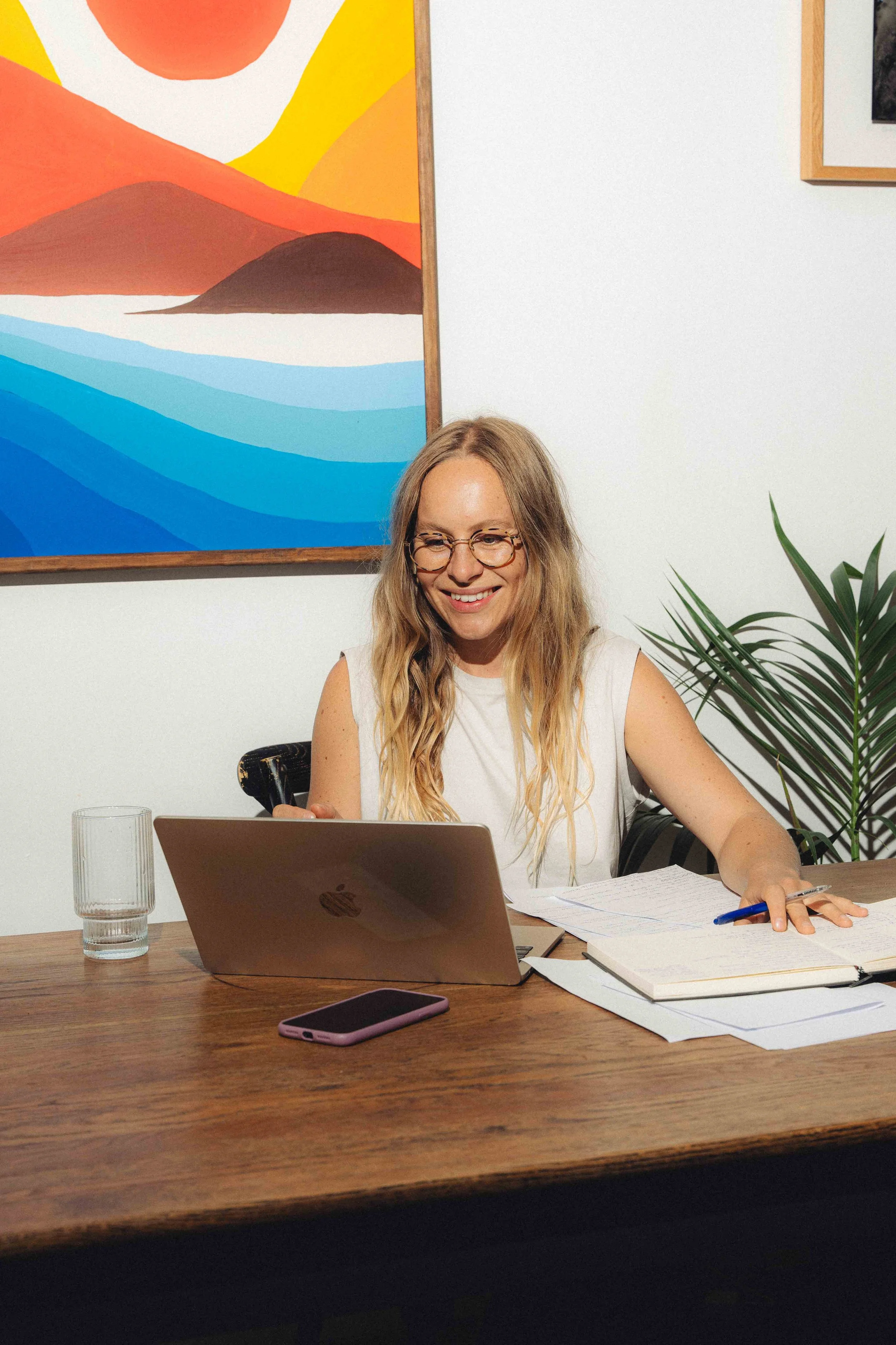 A woman with long blonde hair and glasses smiling while sitting at a desk with a laptop, papers, and a pen in a room with artwork on the wall and a green plant.