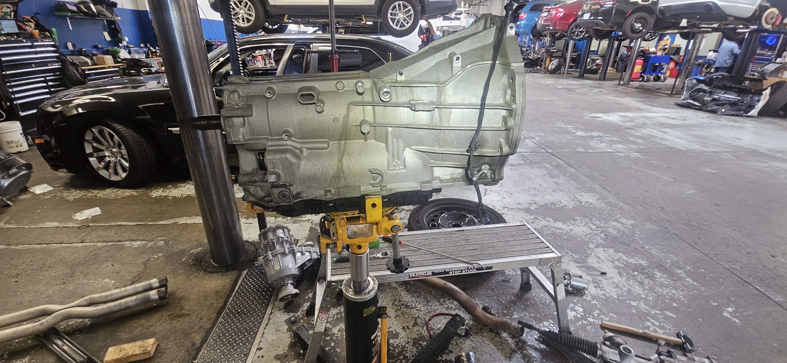 Automotive repair shop with a car engine on a blue stand in the foreground, surrounded by various tools and equipment. Several cars are elevated on lifts in the background.