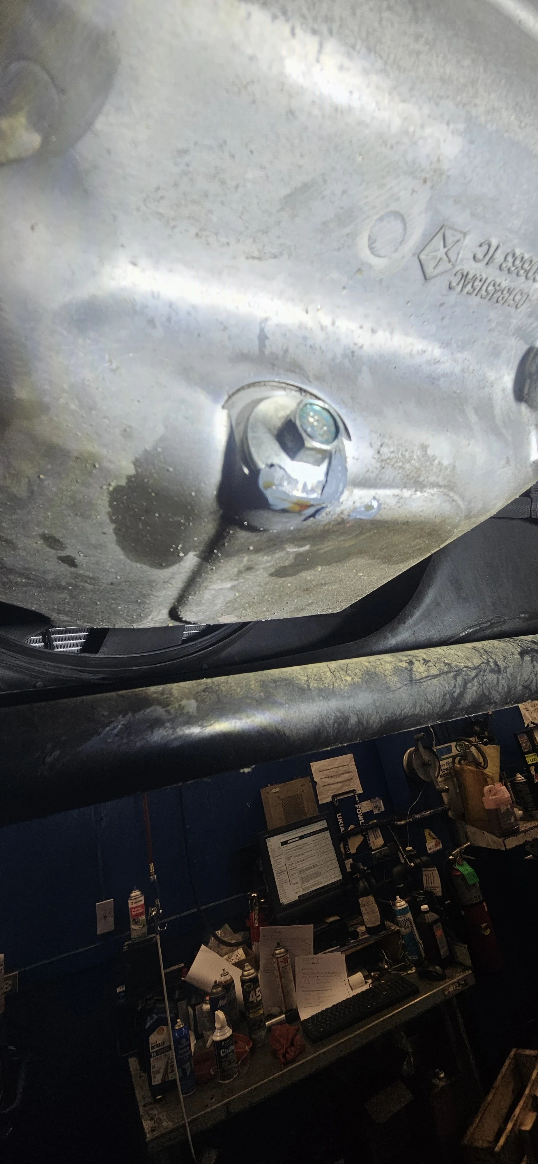 Close-up of a metal part of a machine or vehicle, with a bolt and some marking or engravings, and a workshop or garage setting with tools, papers, and equipment in the background.