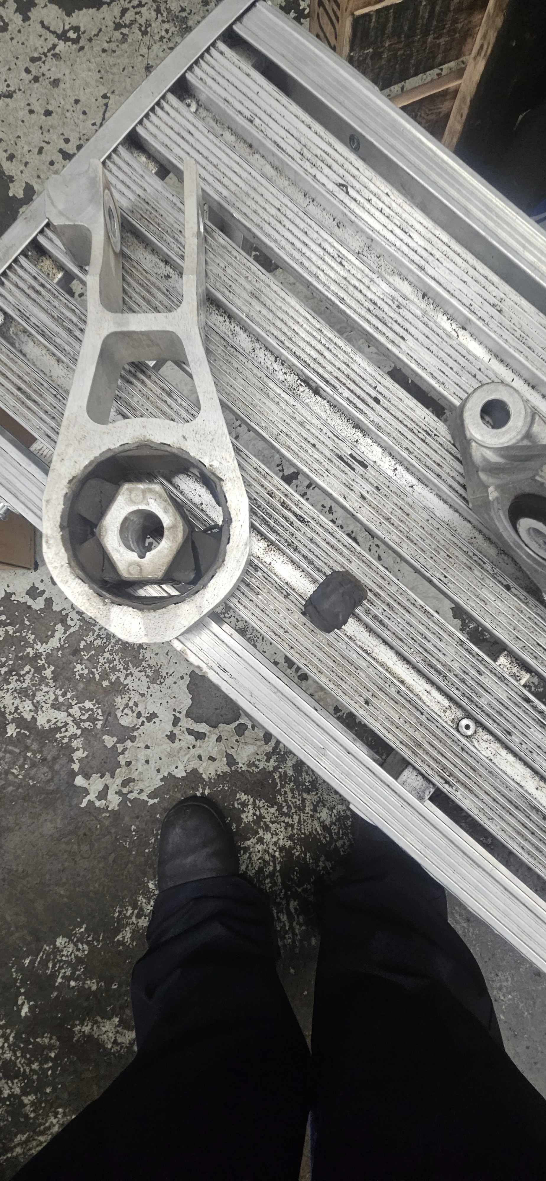 A mechanical part, possibly an engine component, resting on a metal workbench with a person's legs and shoes visible at the bottom of the image. The workbench is in a workshop setting.