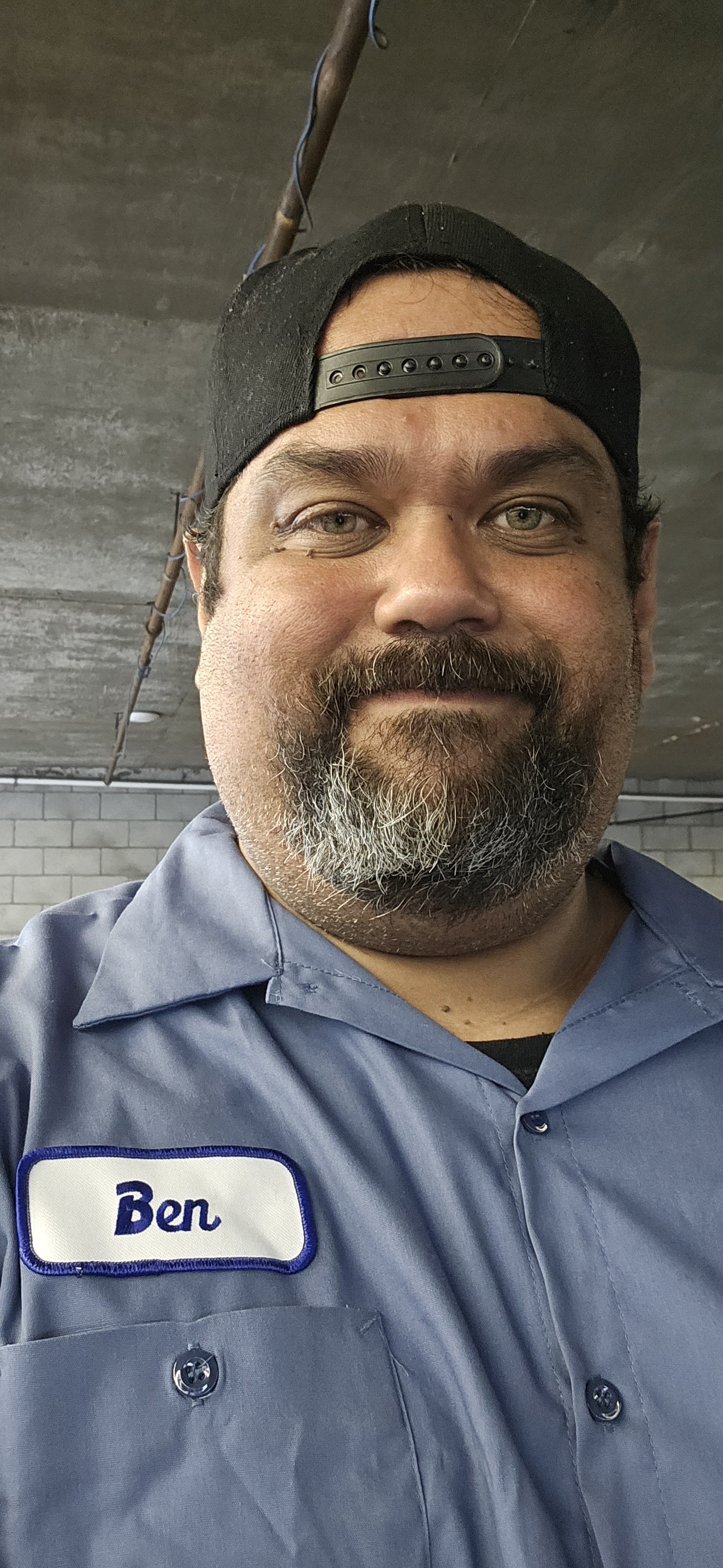 A man with a beard, mustache, and light-colored eyes wearing a black backward cap and a blue work shirt with a name tag that says 'Ben'. He is smiling and appears to be in an industrial or warehouse setting.
