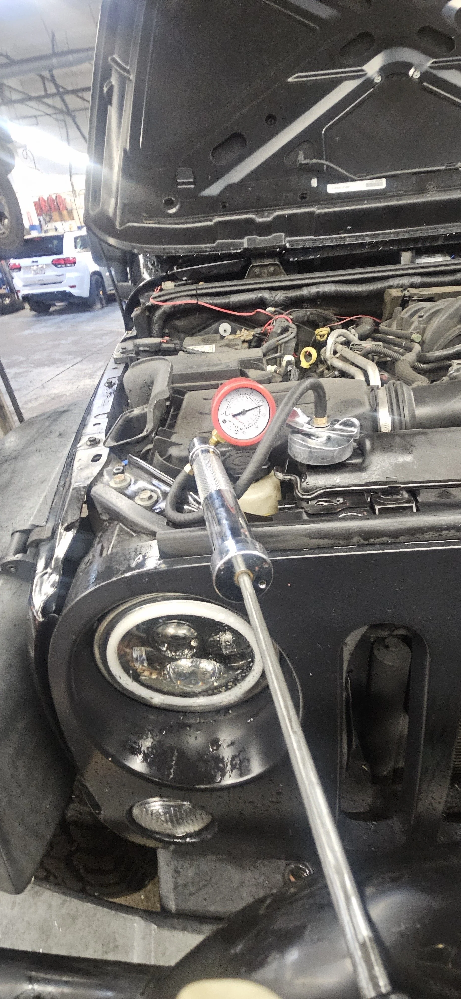 Car with open hood undergoing engine diagnosis or repair, with a mechanic's tool and a pressure gauge visible.