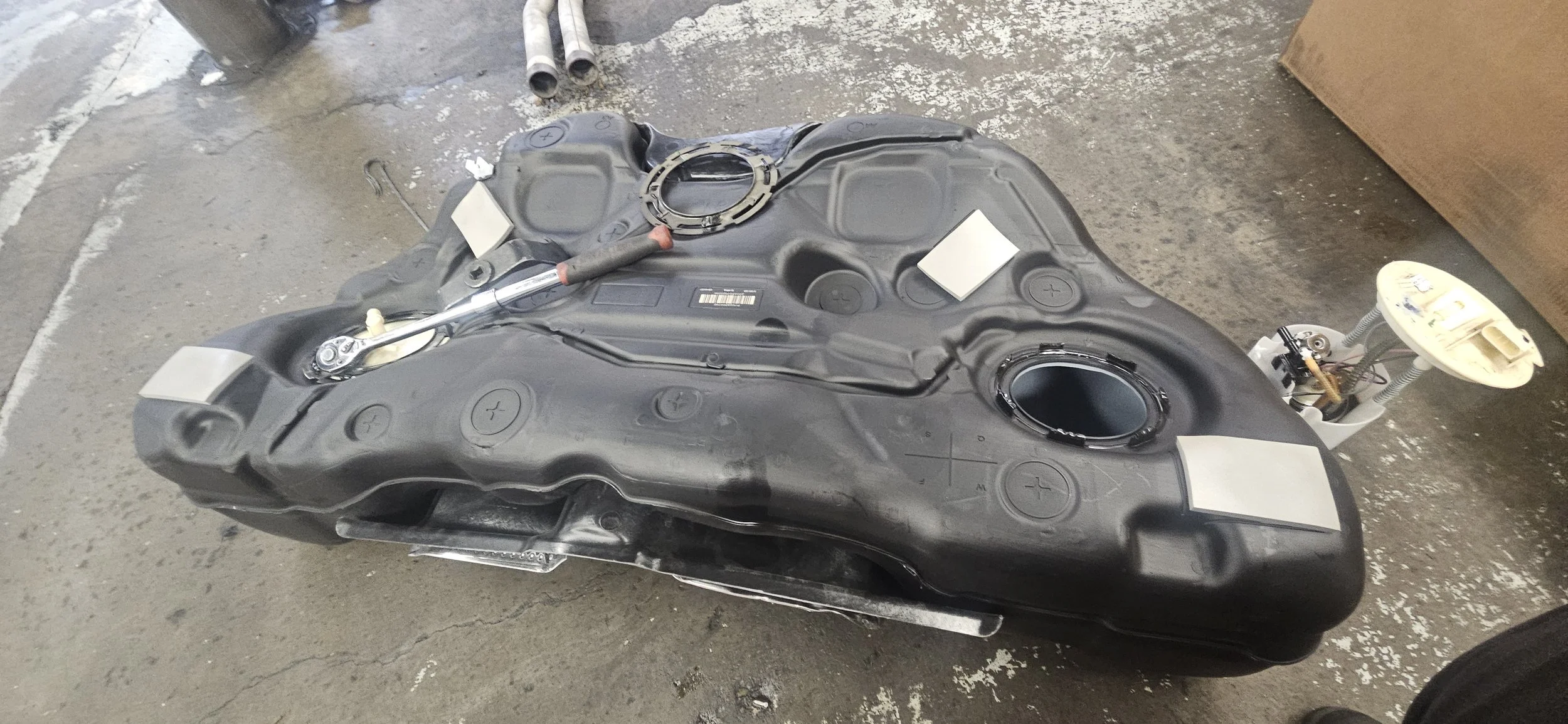 Automotive fuel tank with opening for fuel pump and fuel lines, placed on a rough concrete floor.