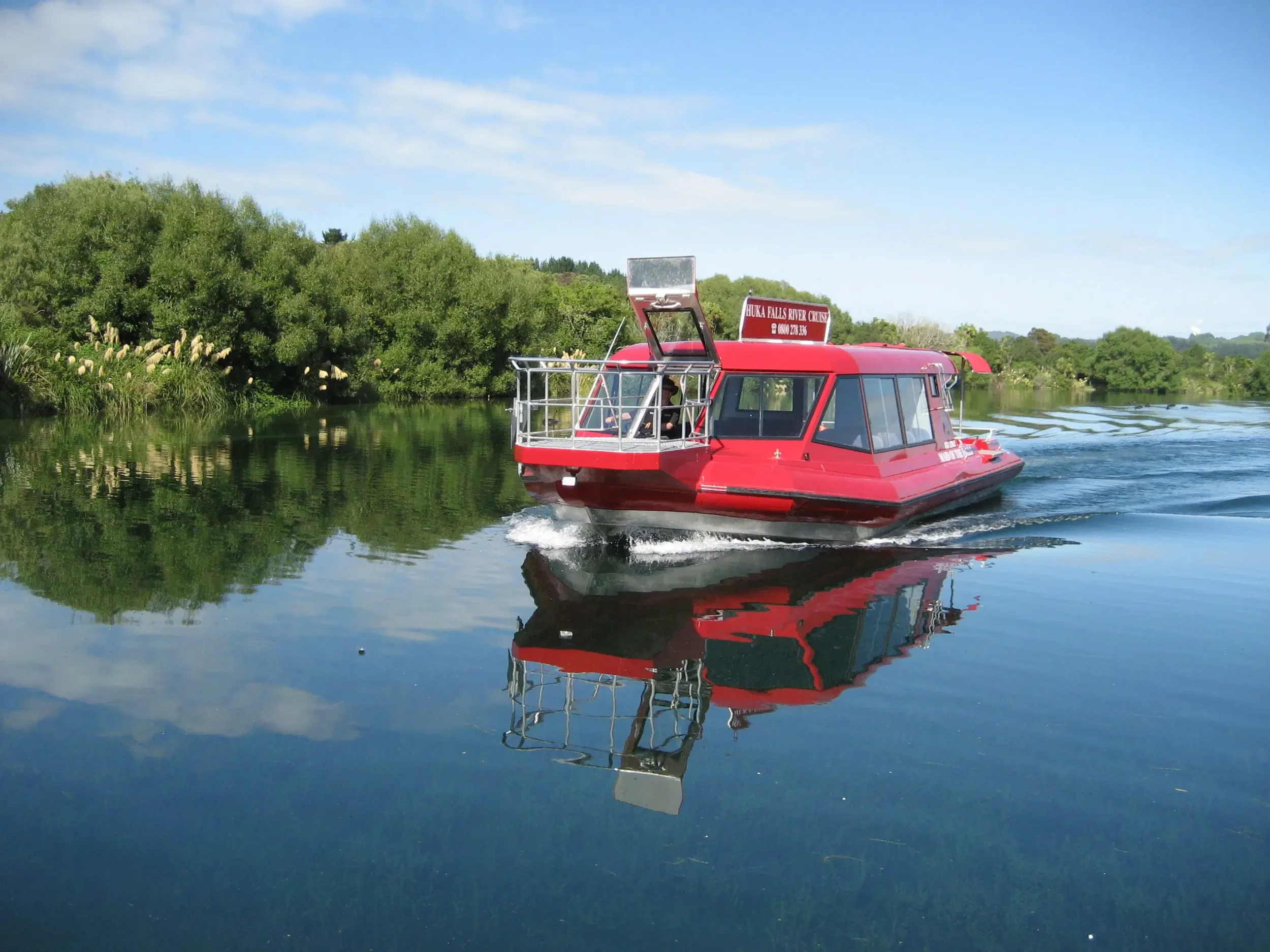 The red boat of the 'Huka Falls River Cruise' on a calm river, surrounded by lush green trees under a partly cloudy sky.