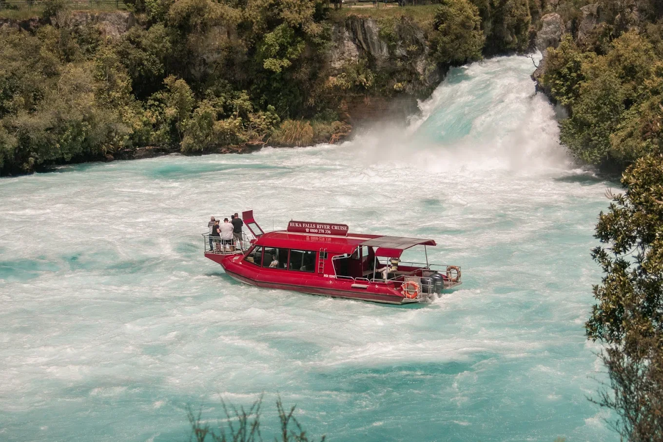 A red boat with a sign that reads 'Huka Falls River Cruise' in the champagne waters below Huka Falls, light blue river surrounded by green foliage.