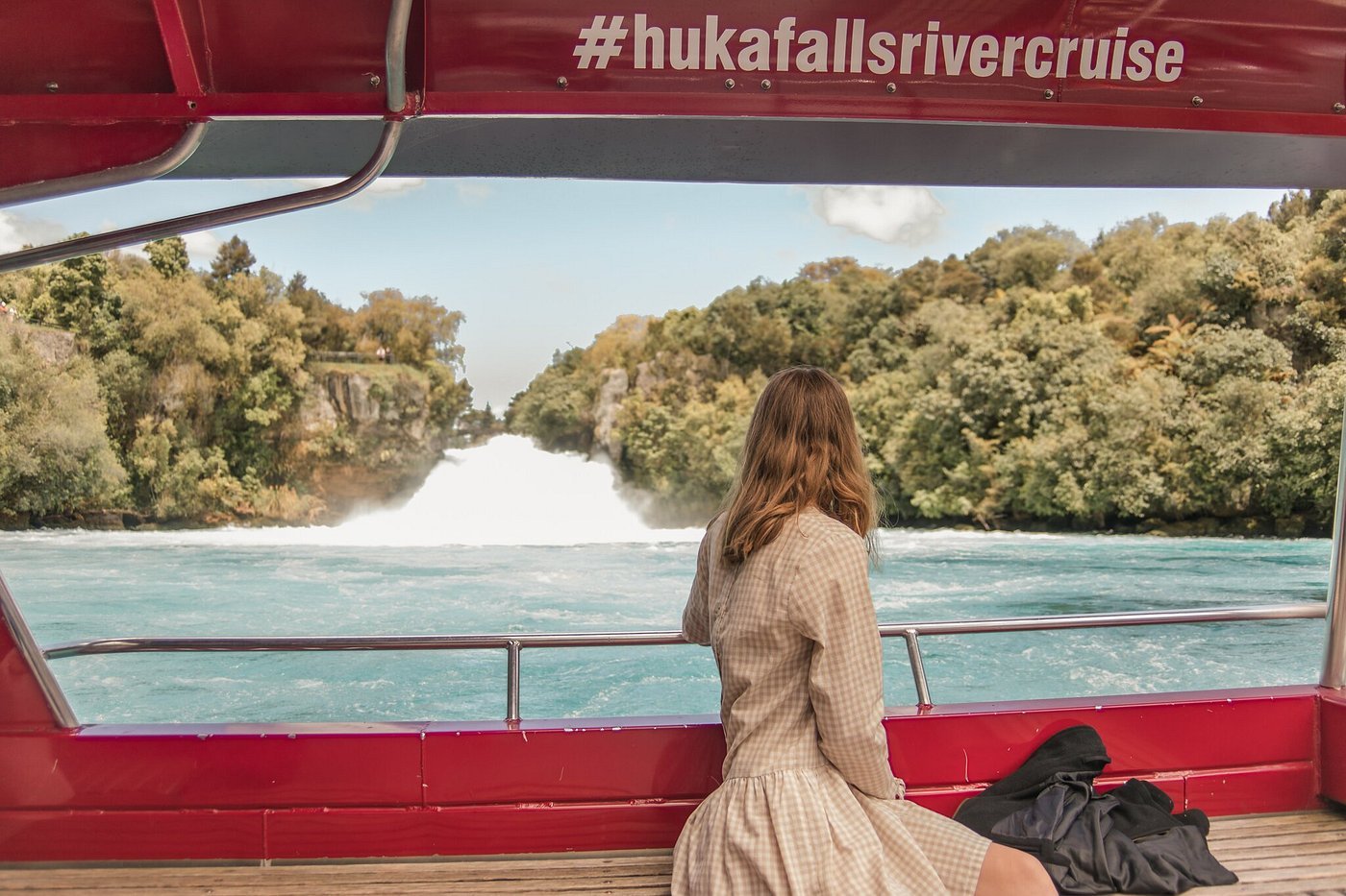 A woman sitting on a boat deck looking at Huka Falls and surrounding greenery during a river cruise.
