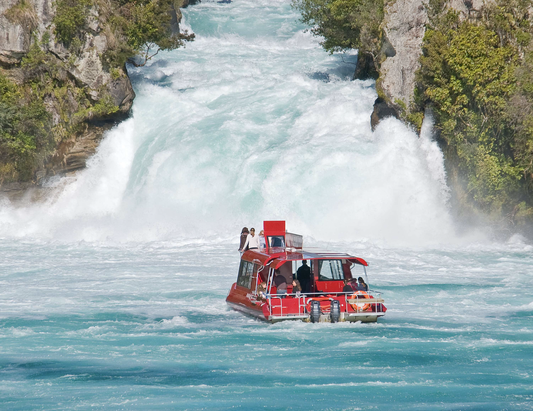 The Huka Falls River Cruise boat with passengers on the Waikato River in front of the Huka Falls surrounded by greenery.
