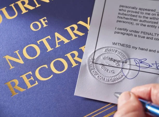 Close-up of a hand signing a document with a pen, with a notary public seal and a notary record book in the background.
