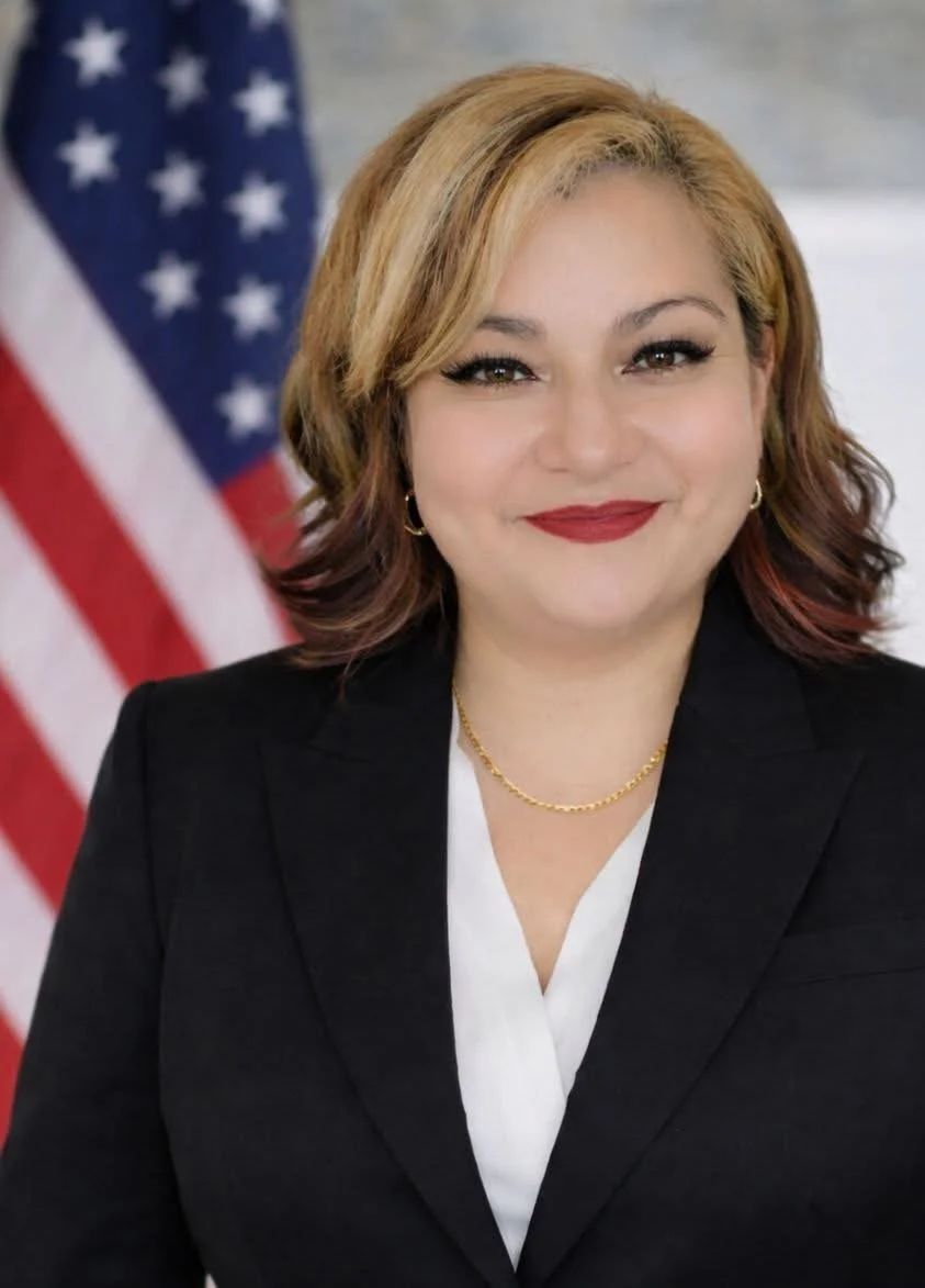 A woman with blonde and reddish hair wearing a black blazer, white blouse, gold necklace, and gold hoop earrings, standing in front of an American flag.