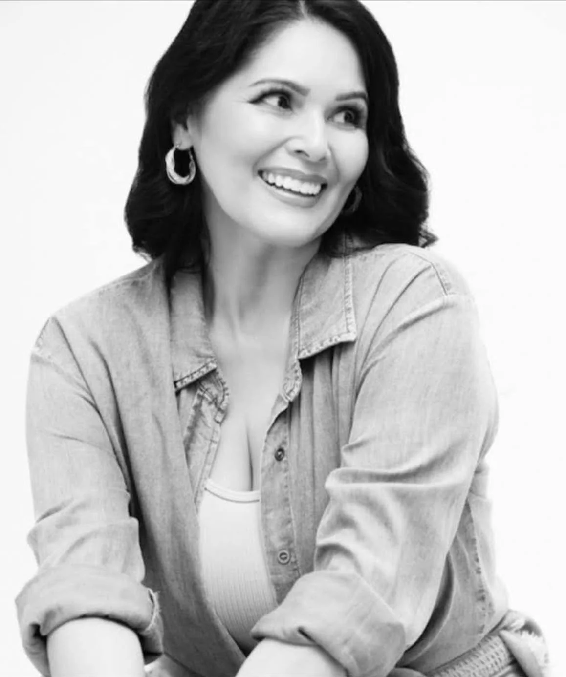 Black and white photo of a smiling woman with dark wavy hair, wearing hoop earrings and a button-up denim shirt, looking to her right with a cheerful expression.