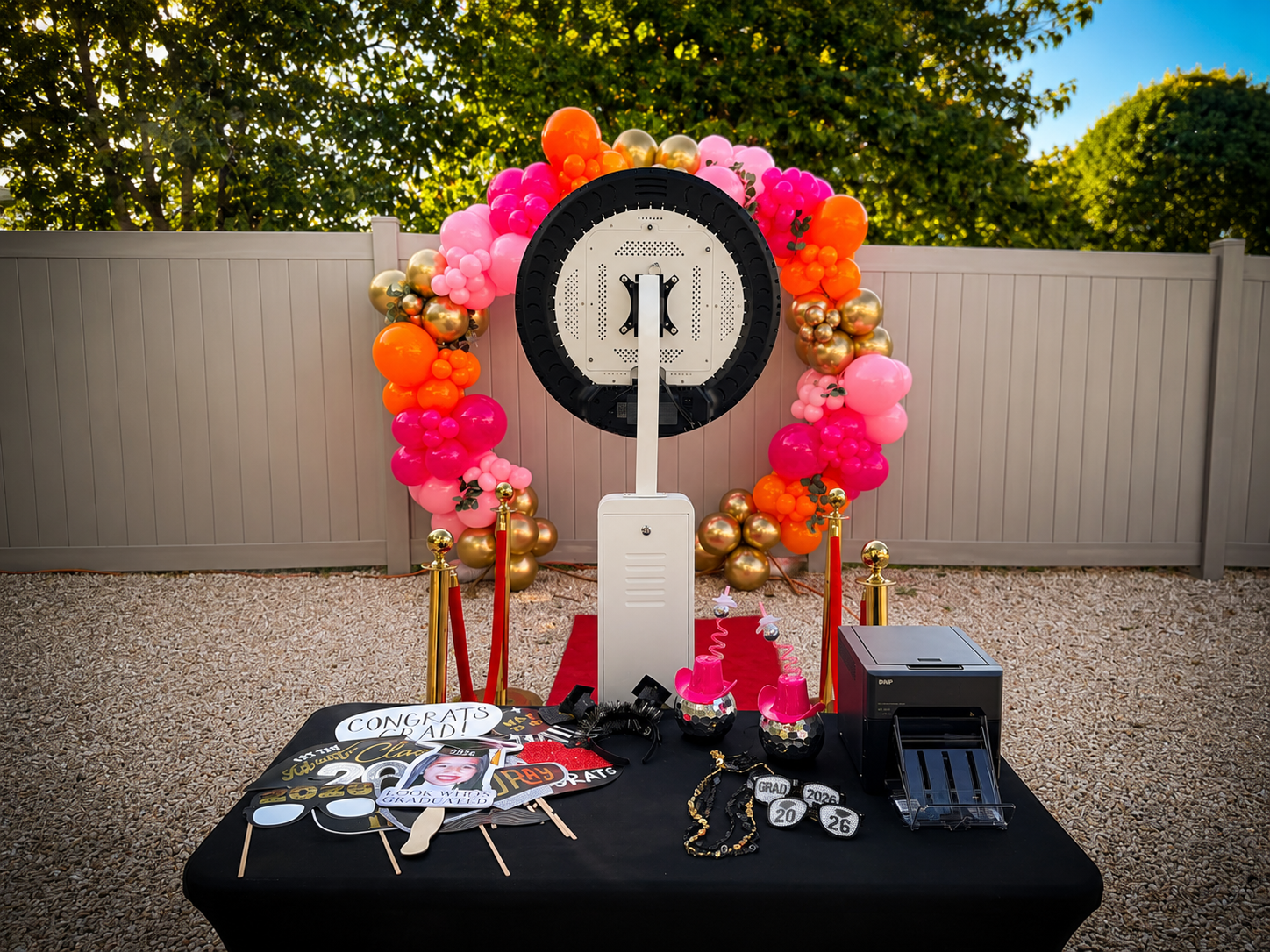 Photo of a graduation celebration setup with a backdrop decorated with pink, orange, and gold balloons, a table with congratulatory signs, photo props, and graduation accessories, outdoors on a sunny day.