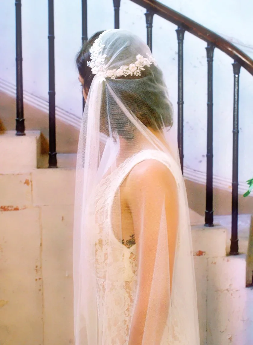 A woman in a wedding dress with a veil and floral headpiece stands near a staircase with black iron railing, facing away.