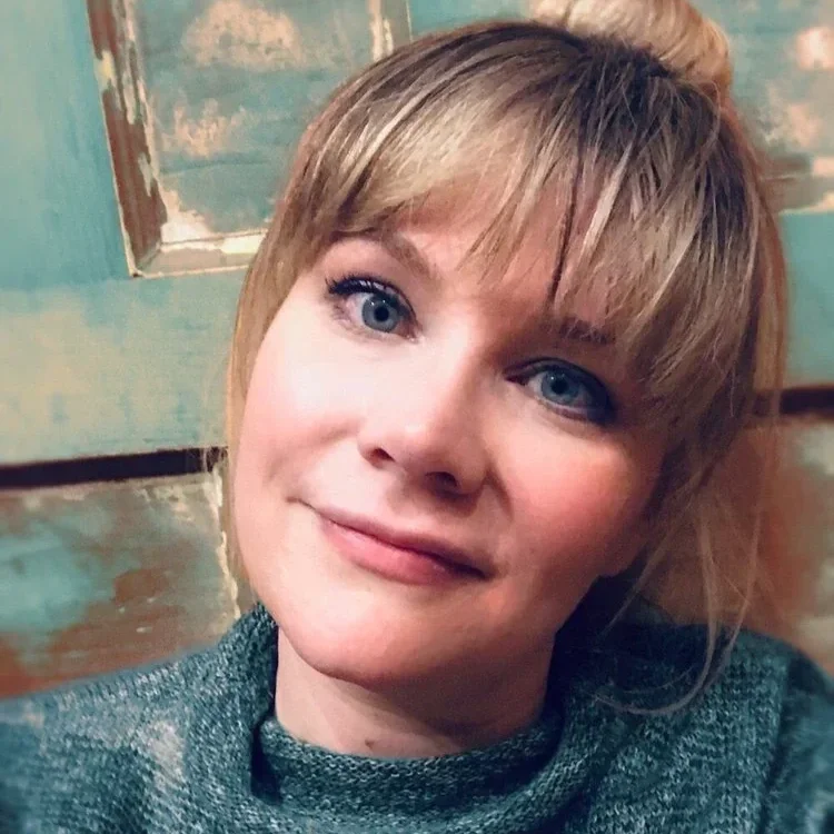 A woman with short blonde hair and blue eyes smiling at the camera, wearing a gray sweater, in front of a worn, painted wooden wall.