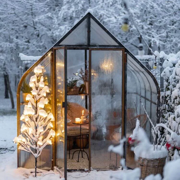 Snow-covered greenhouse with warm lights inside, decorated with a glowing tree and a small candlelit table, surrounded by snow-covered garden and pink flowers in a basket.